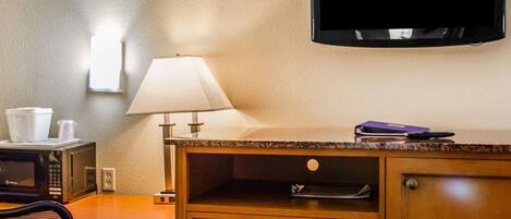 A hotel room with a wooden desk, a chair, a television mounted on the wall, a lamp, and a microwave on the counter.