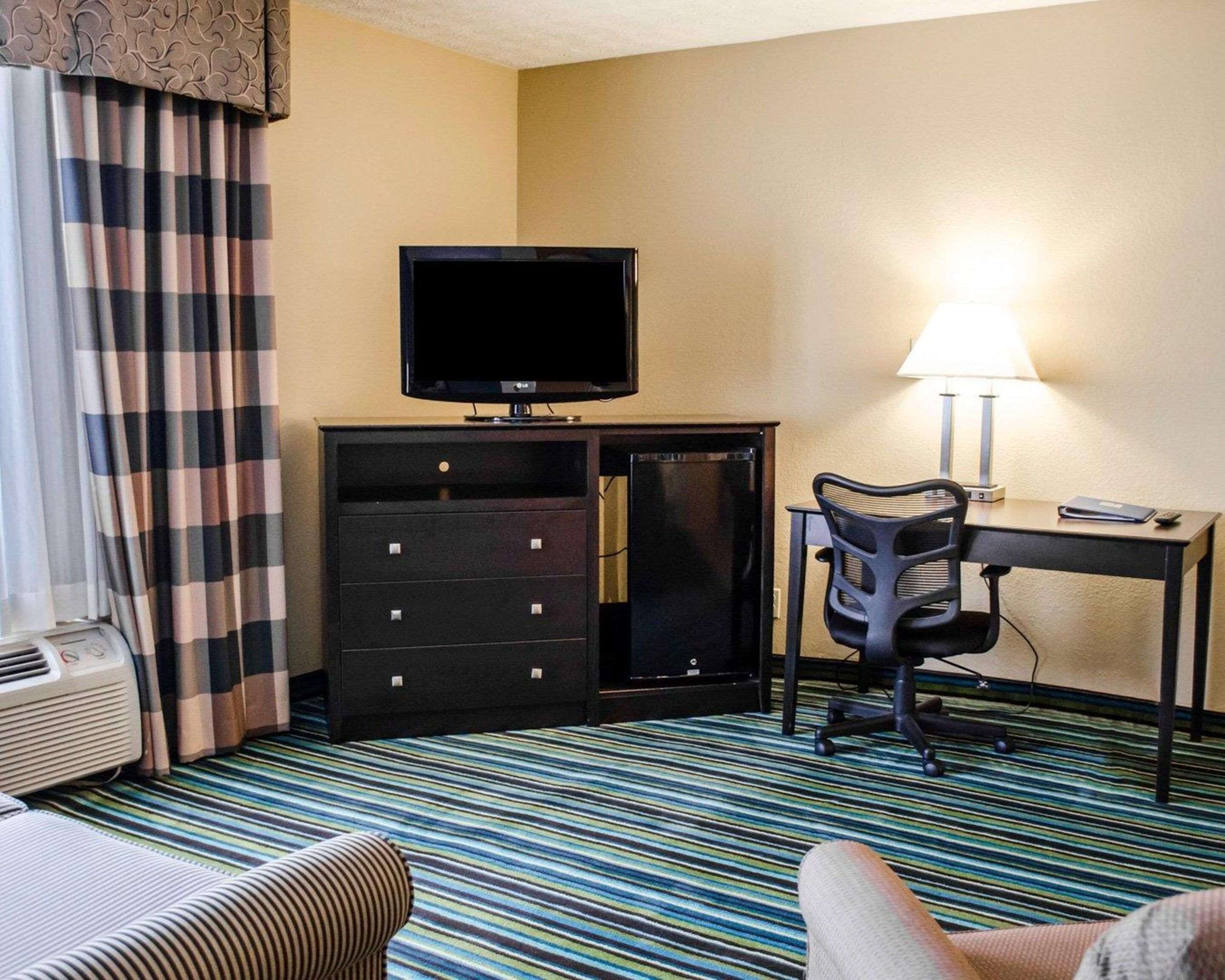 Habitación de hotel con televisión, escritorio con lámpara, una silla, cómoda y alfombra a rayas.