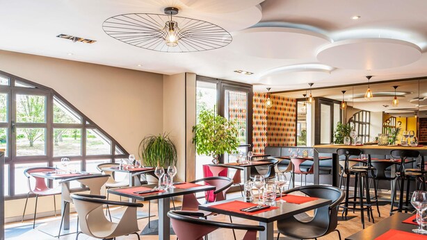 Interior moderno de restaurante con techos altos, ventanas grandes y una combinación de sillas rojas y negras.
