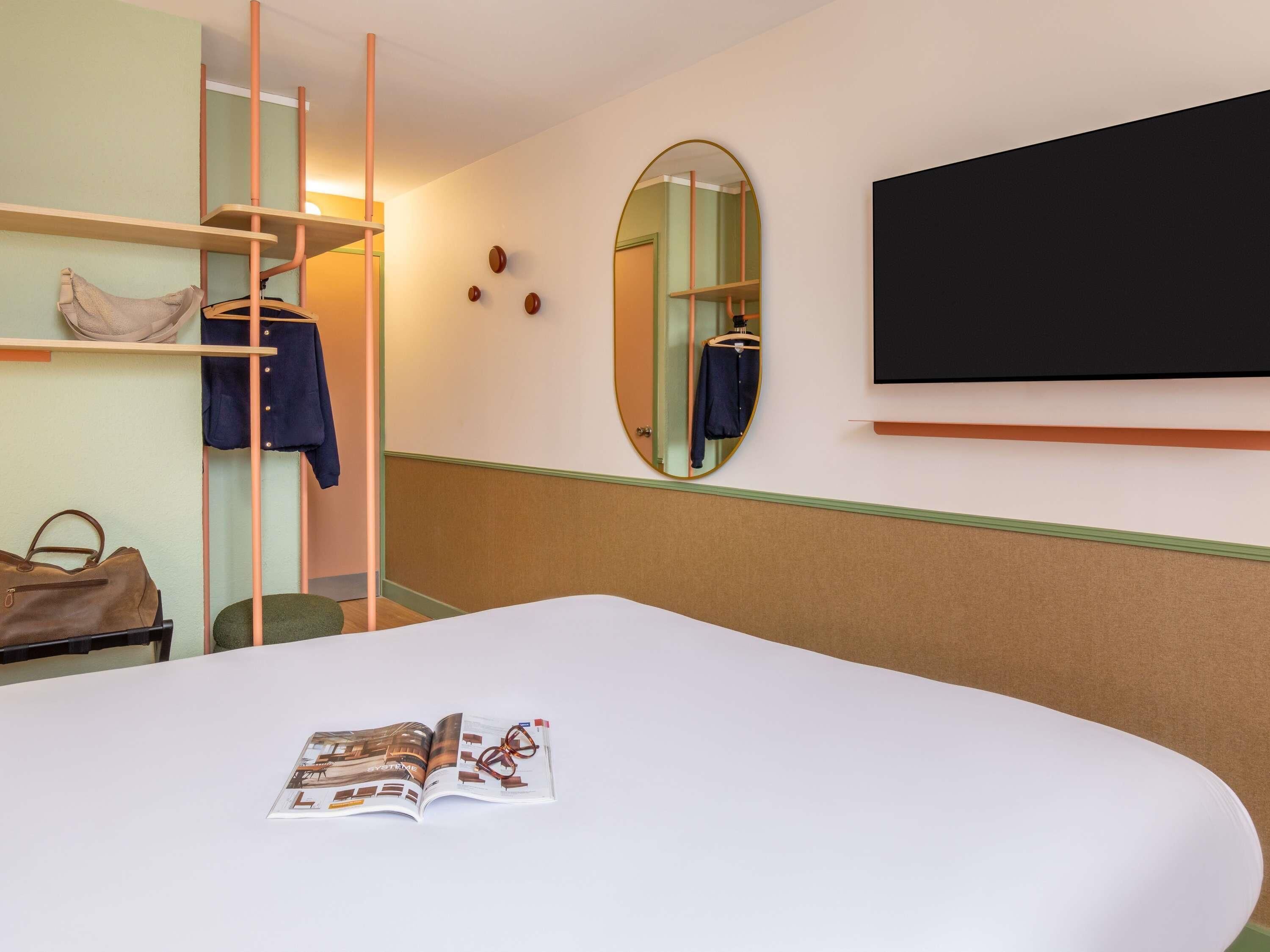 Une chambre d’h ôtel avec un lit, un magazine, un miroir, une télévision et un porte-manteau.