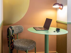A laptop on a round table with a chair beside it, a wall-mounted lamp, and a partial view of a room with a curved wall. - ibis Styles Reims Centre Cathédrale (Reims)