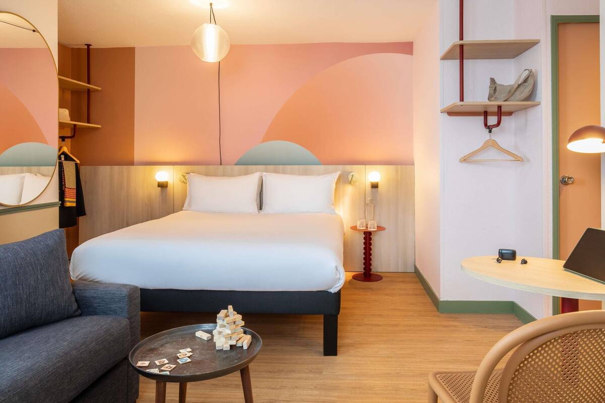 A hotel room with a bed, a sofa, a small table with wooden blocks, a desk with a laptop, and a hanging coat rack.