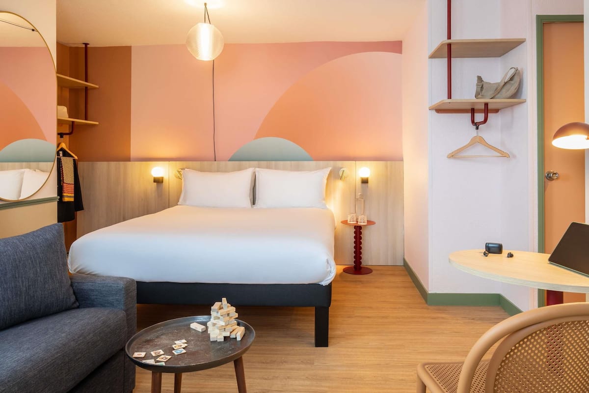 Quarto de hotel com cama, sofá, pequena mesa com blocos de madeira, escrivaninha com laptop e cabideiro.