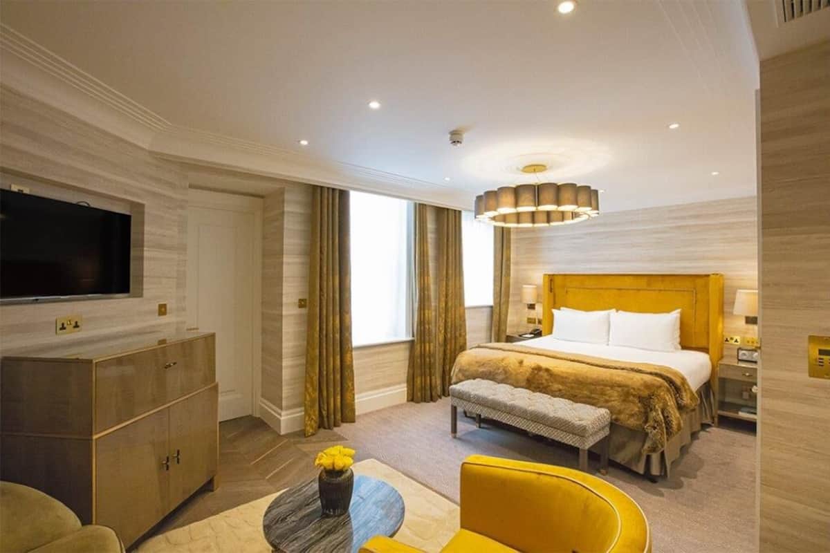 A hotel room with a large bed, a television on the wall, a desk with a lamp, and a window with curtains.