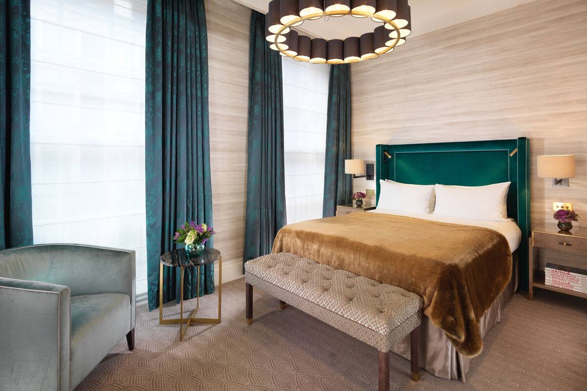 A hotel room with a large bed, a bench, a chair, a round chandelier, and a small table with a vase of flowers.