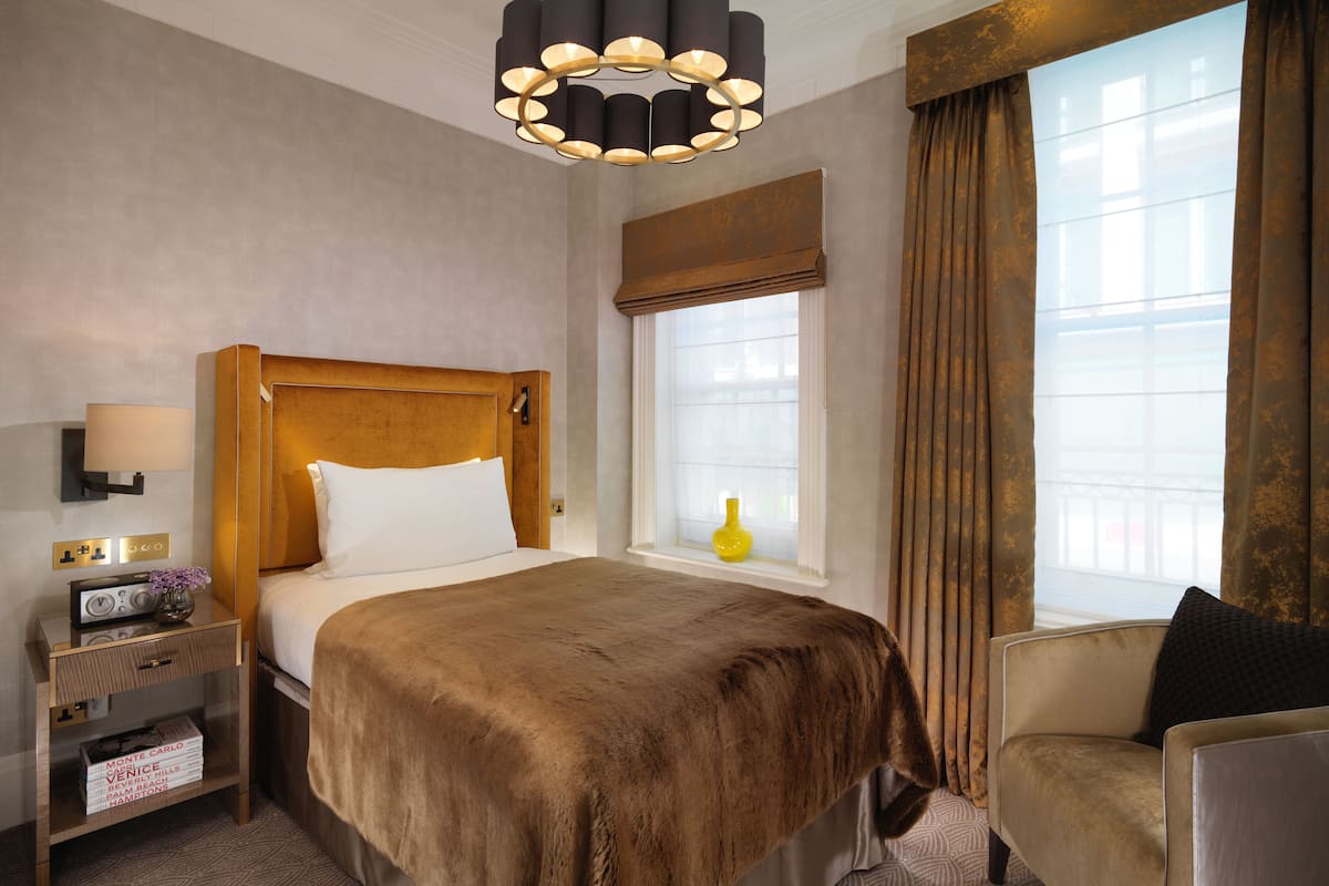 A hotel room with a large bed, a bedside table with a lamp, a small table with a vase, and a window with curtains.