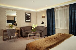Ein Hotelzimmer mit Bett, Schreibtisch, Sofa, Sessel, Couchtisch und einem großen Fenster mit Vorhängen.