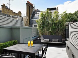 A rooftop patio with a table and chairs, two glasses of orange juice, and a seating area with cushions.