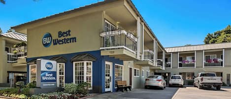A Best Western hotel with a blue and beige facade, a balcony with potted plants, and a parking area with cars.