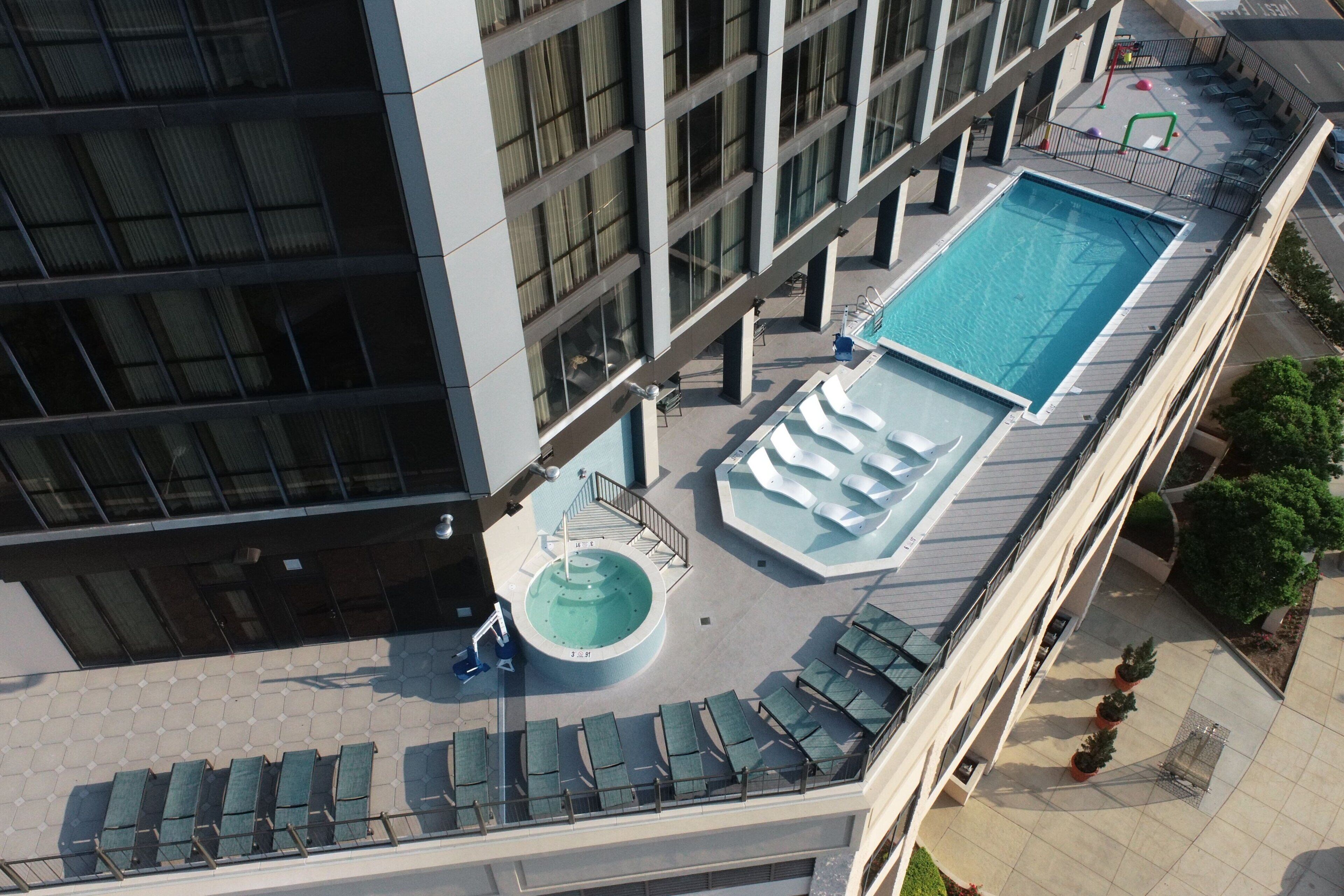 An aerial view of a hotel rooftop with a swimming pool, sun loungers, and a circular hot tub.