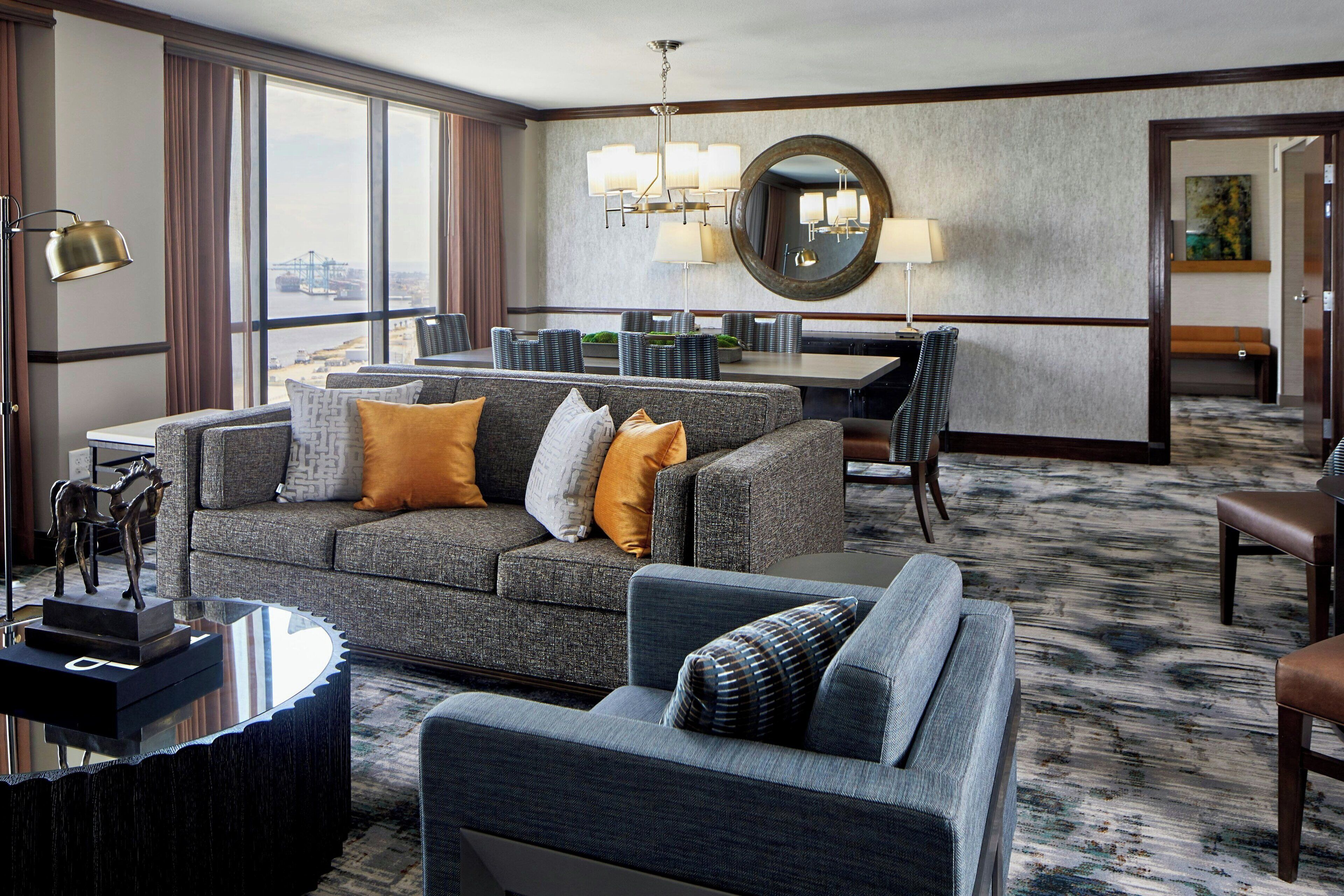 A modern hotel room with a grey sofa set, a dining table, and a large window offering a city view.