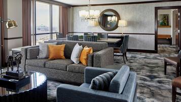 A modern hotel room with a grey sofa set, a dining table, and a large window offering a city view.
