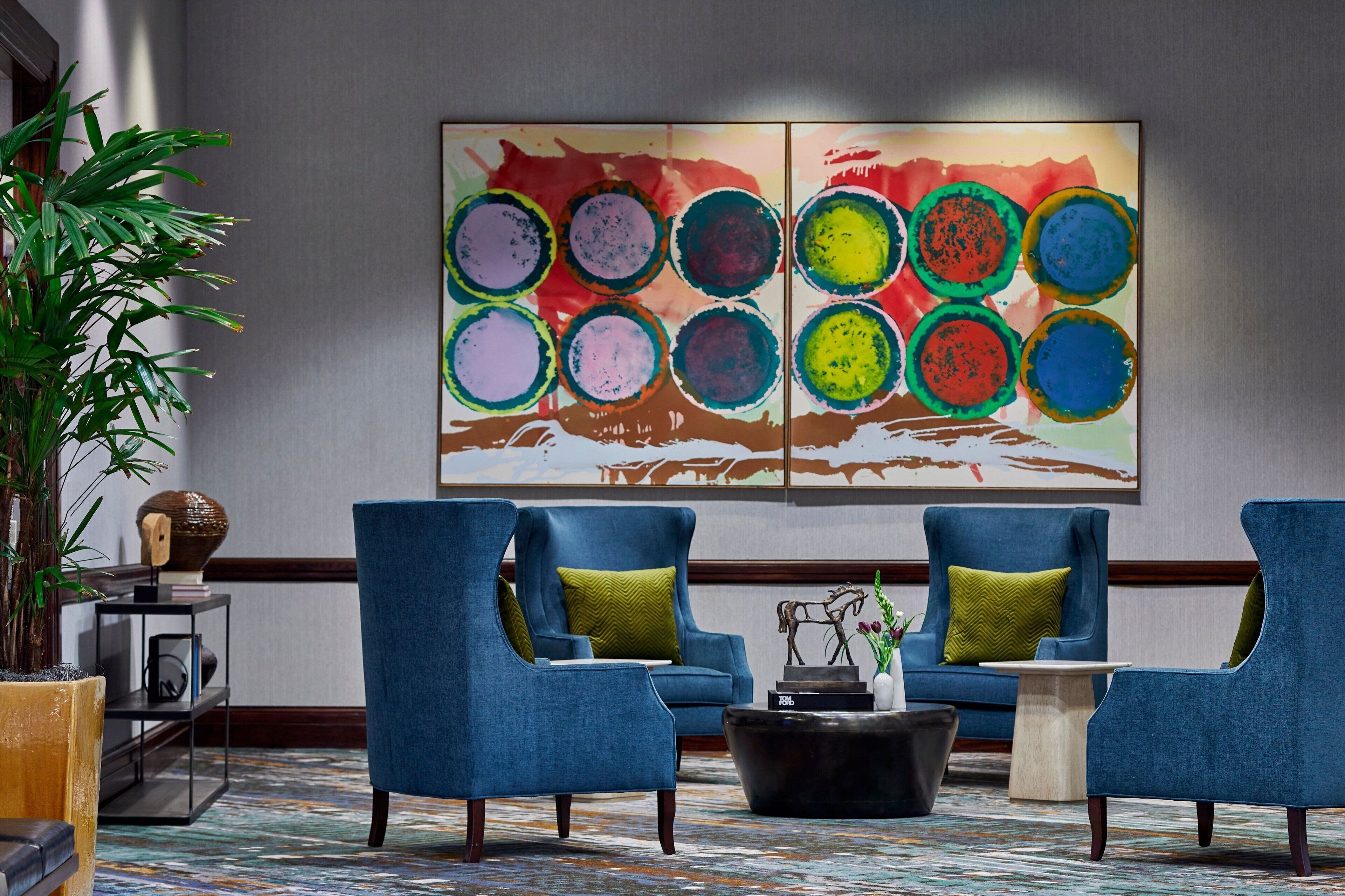 A lobby with two blue armchairs, a large abstract painting, a potted plant, and a small table with a vase.