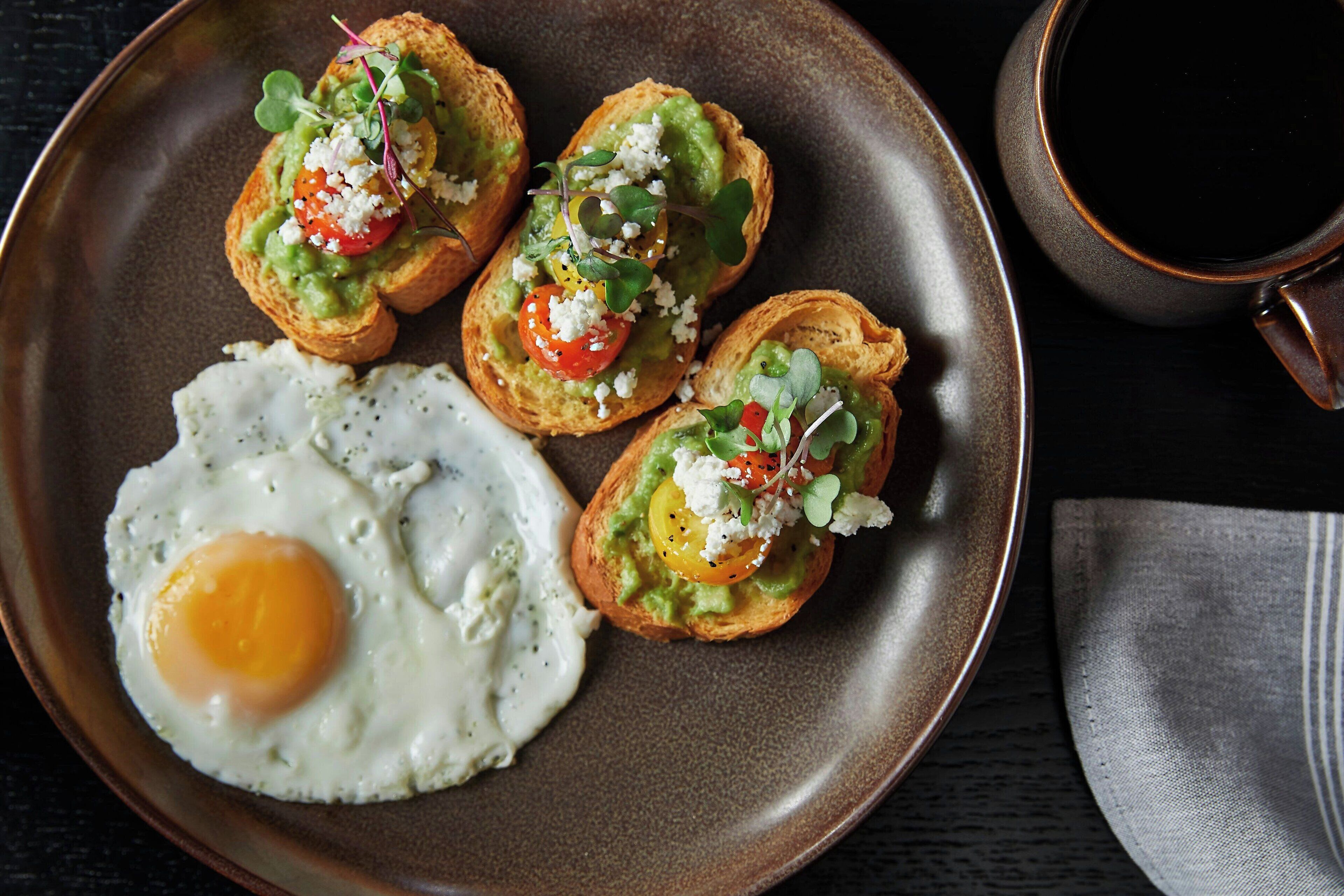 Un plato con cuatro tostadas de aguacate cubiertas de tomates, queso y verduras, junto a un huevo frito.