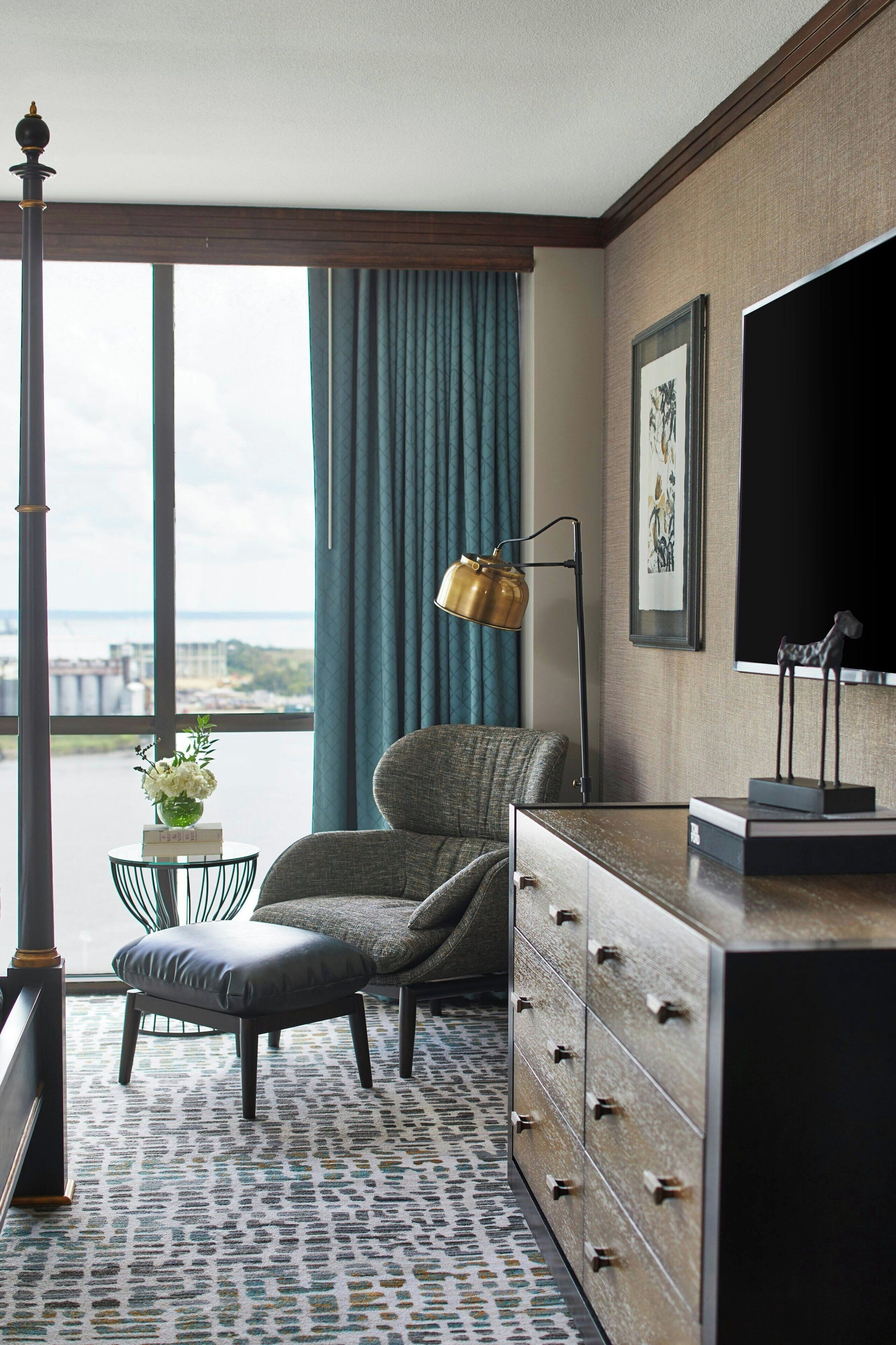 Une chambre d’hôtel dotée d’une grande fenêtre, offrant une vue sur la ville, avec un bureau et une lampe, une chaise, une commode et un tableau encadré au mur.