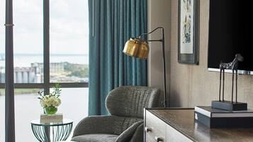 Une chambre d’hôtel dotée d’une grande fenêtre, offrant une vue sur la ville, avec un bureau et une lampe, une chaise, une commode et un tableau encadré au mur.