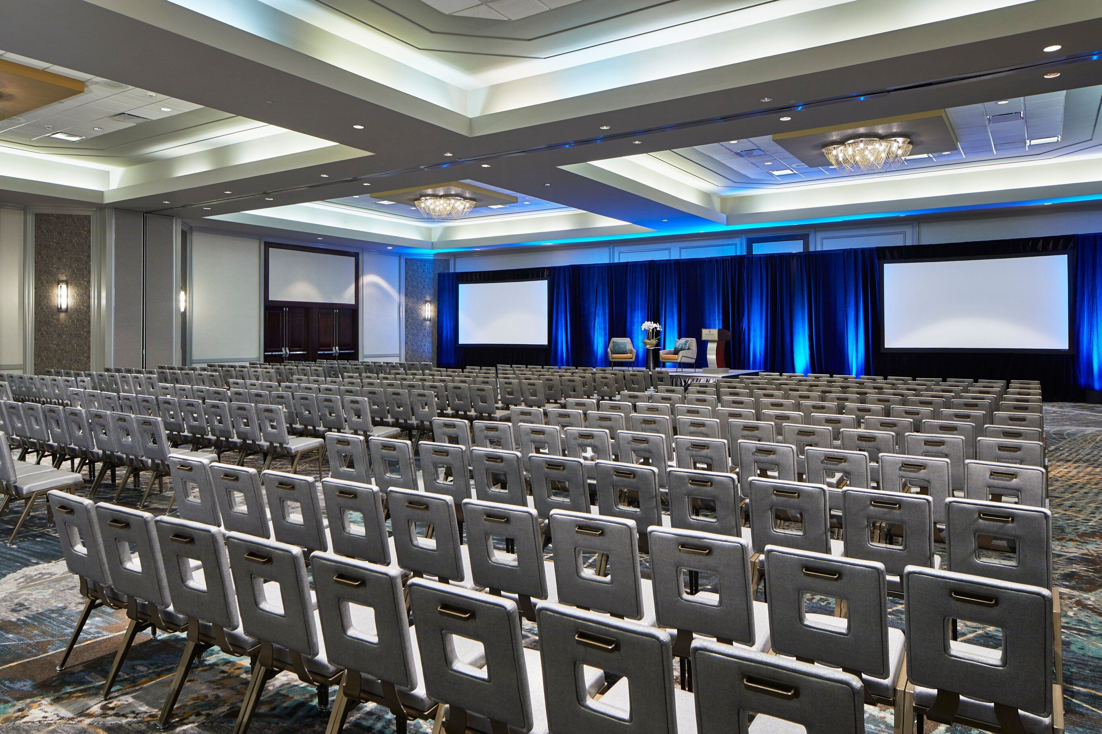 Une salle de conférence avec des rangées de chaises, un grand écran et une scène surmontée d’un rideau bleu.