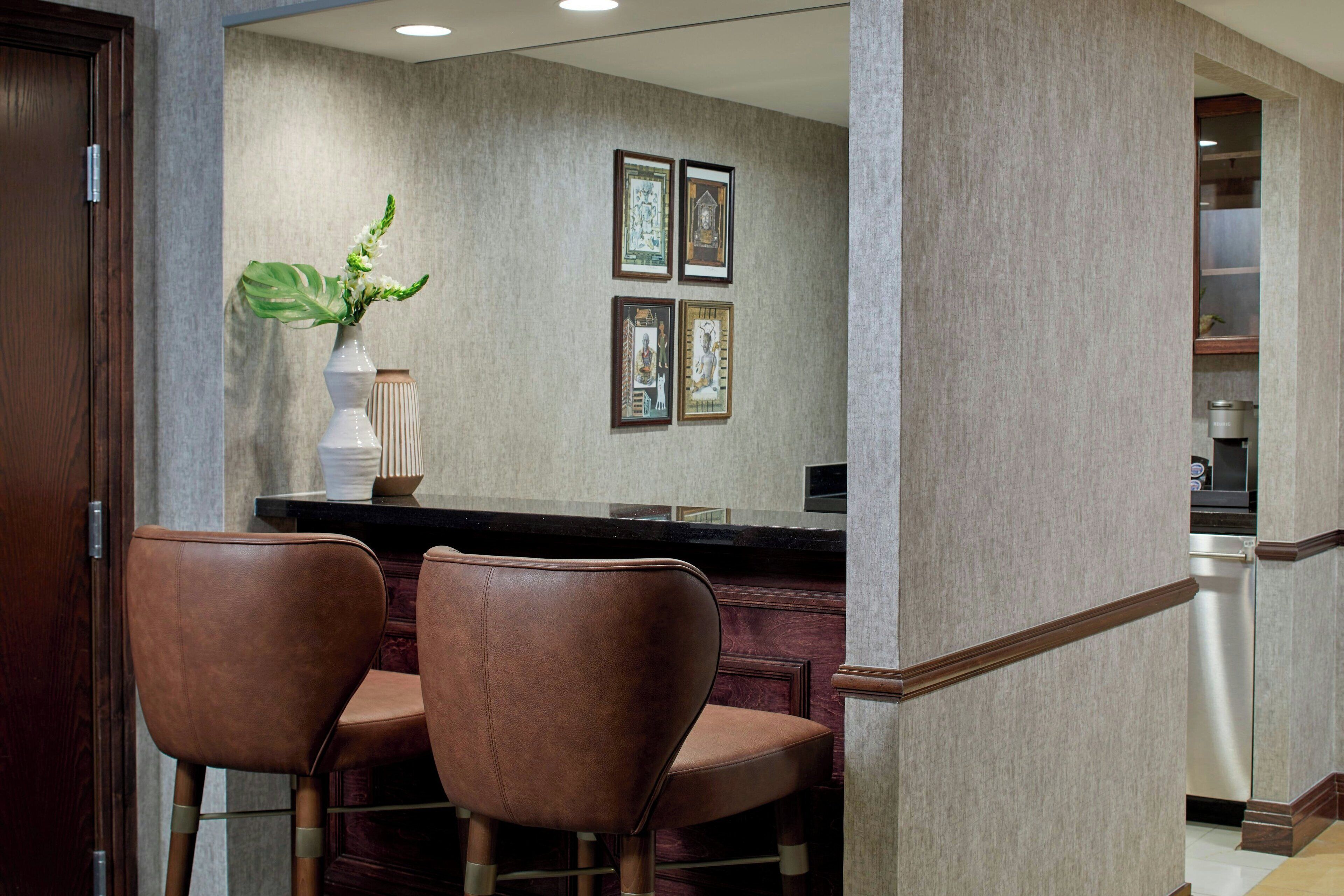 A hotel bar area with two brown leather chairs, a vase with green foliage, and framed pictures on the wall.