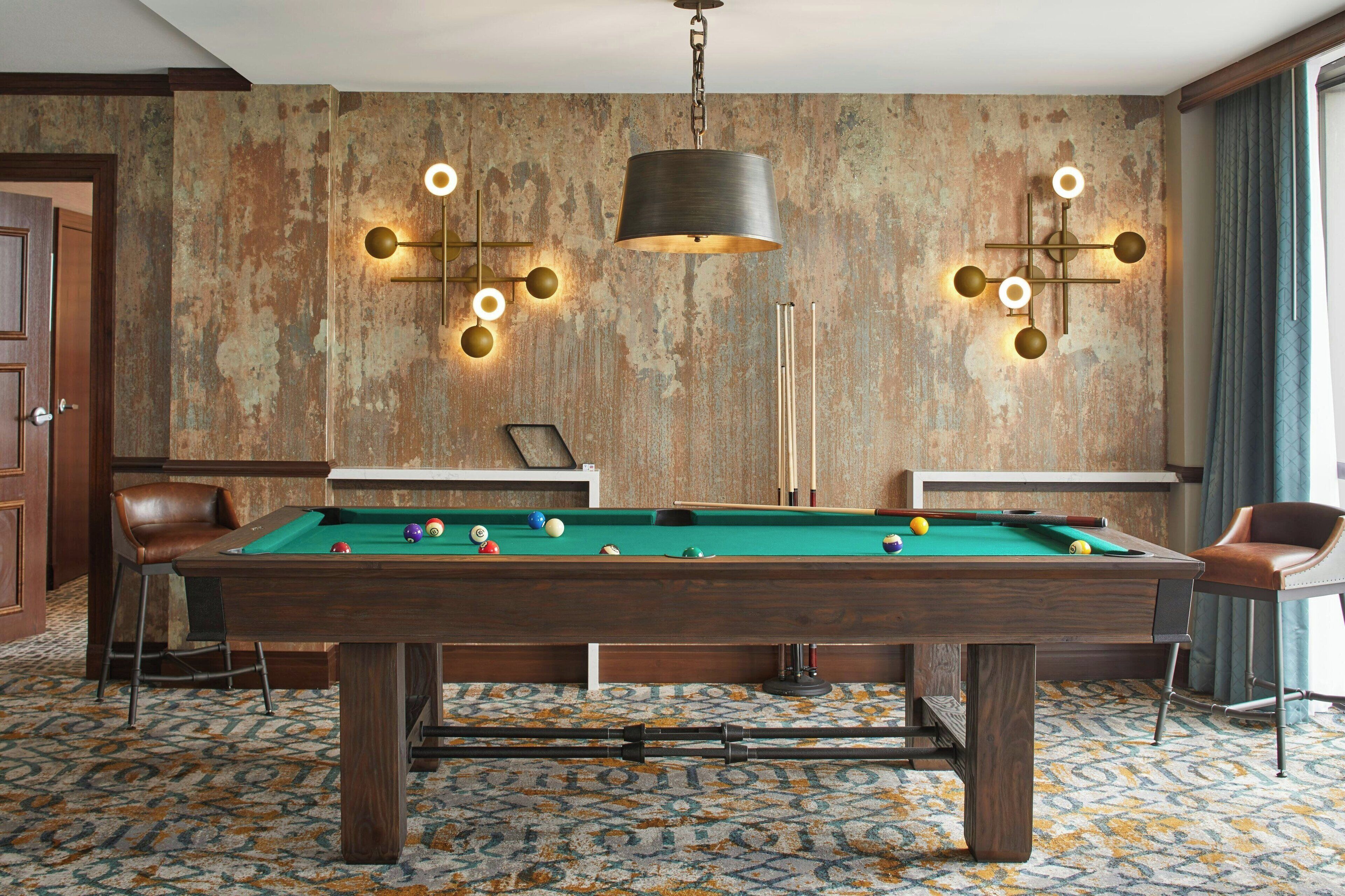 Una mesa de billar con bolas, dos taburetes de bar, una lámpara de pared y una alfombra estampada.