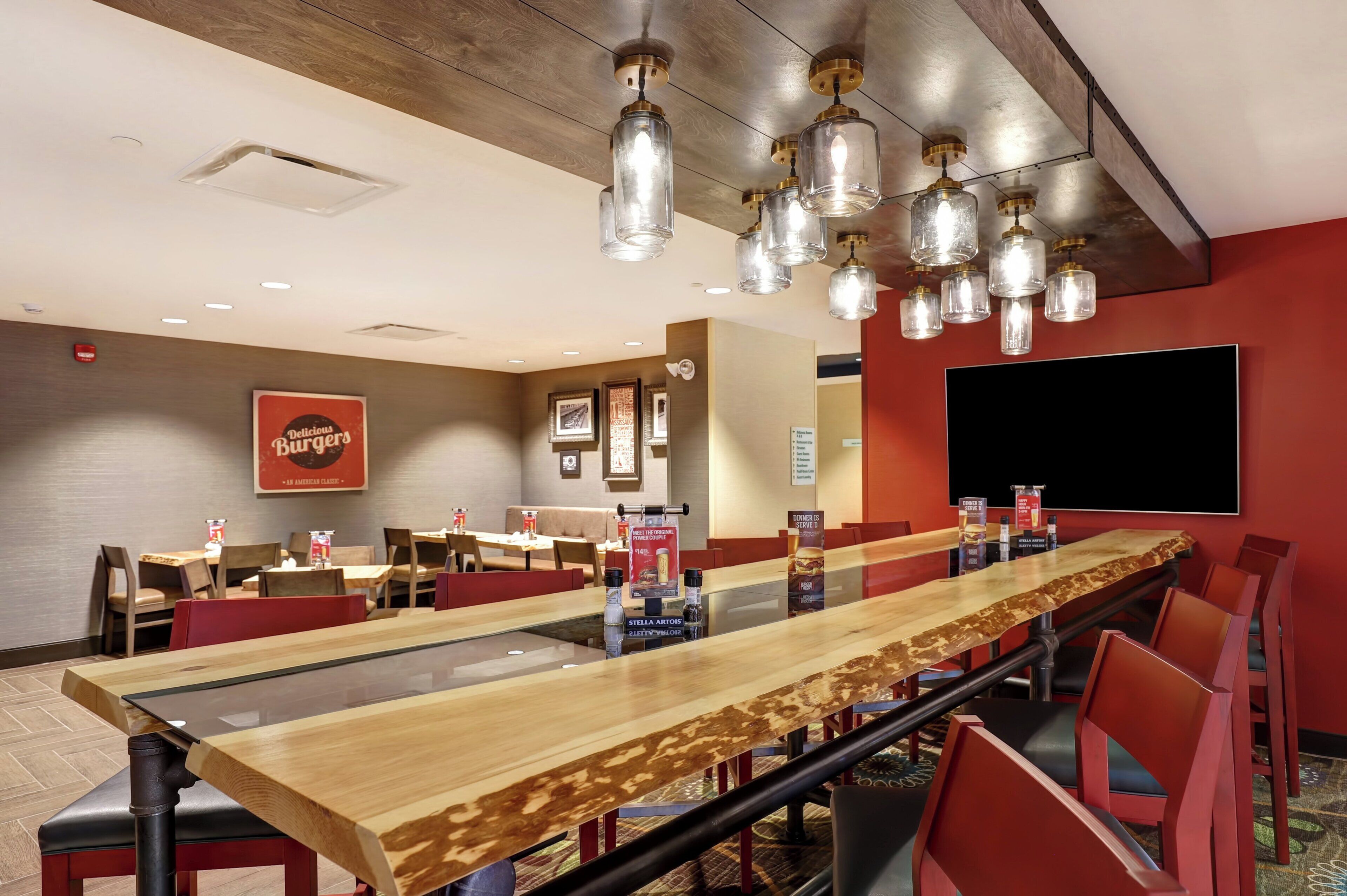 A restaurant with long communal tables, red chairs, and hanging pendant lights.