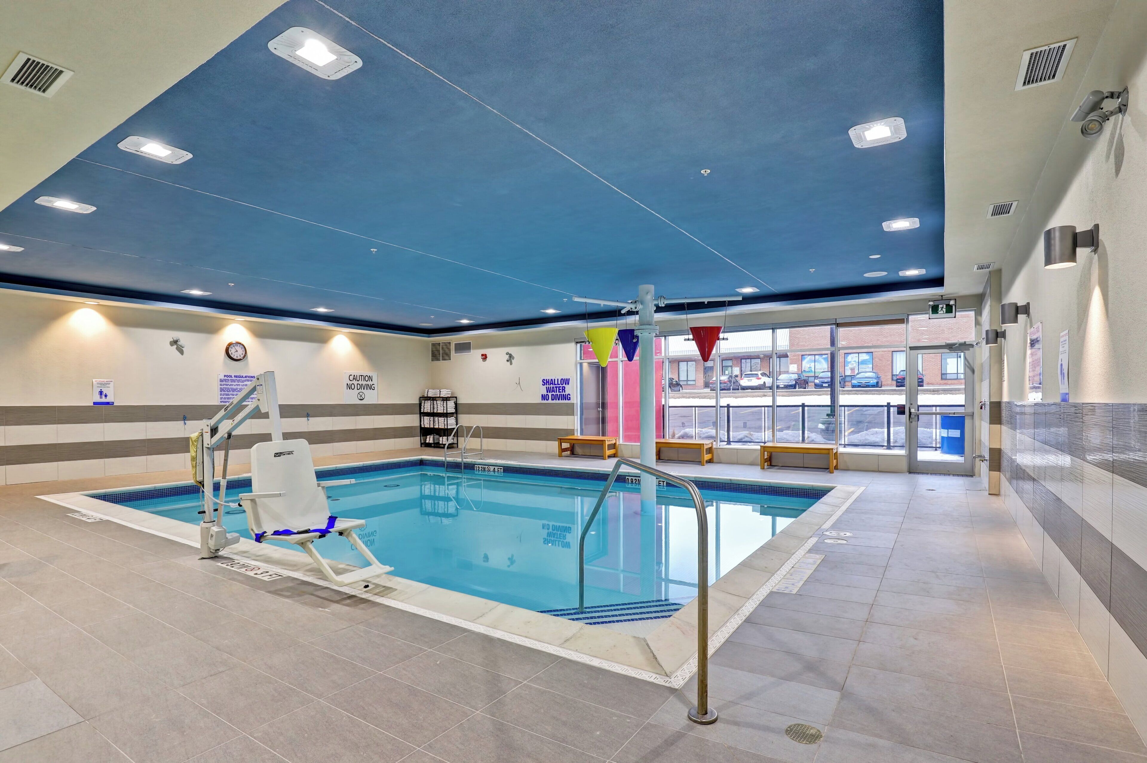 Une piscine intérieure avec un plafond bleu, des panneaux de sécurité et une vue sur l’extérieur.