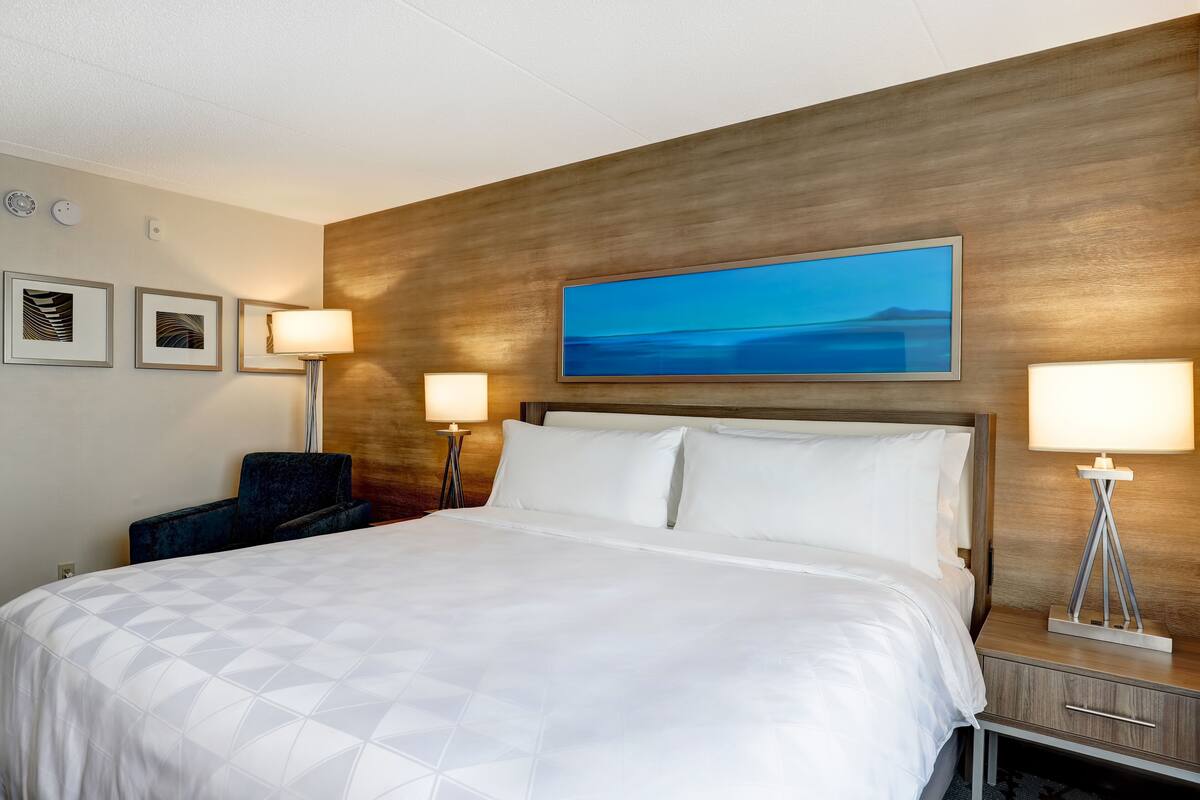 Une chambre d’hôtel avec un grand lit, deux tables de chevet avec des lampes, une chaise et des œuvres d’art encadrées accrochées au mur.