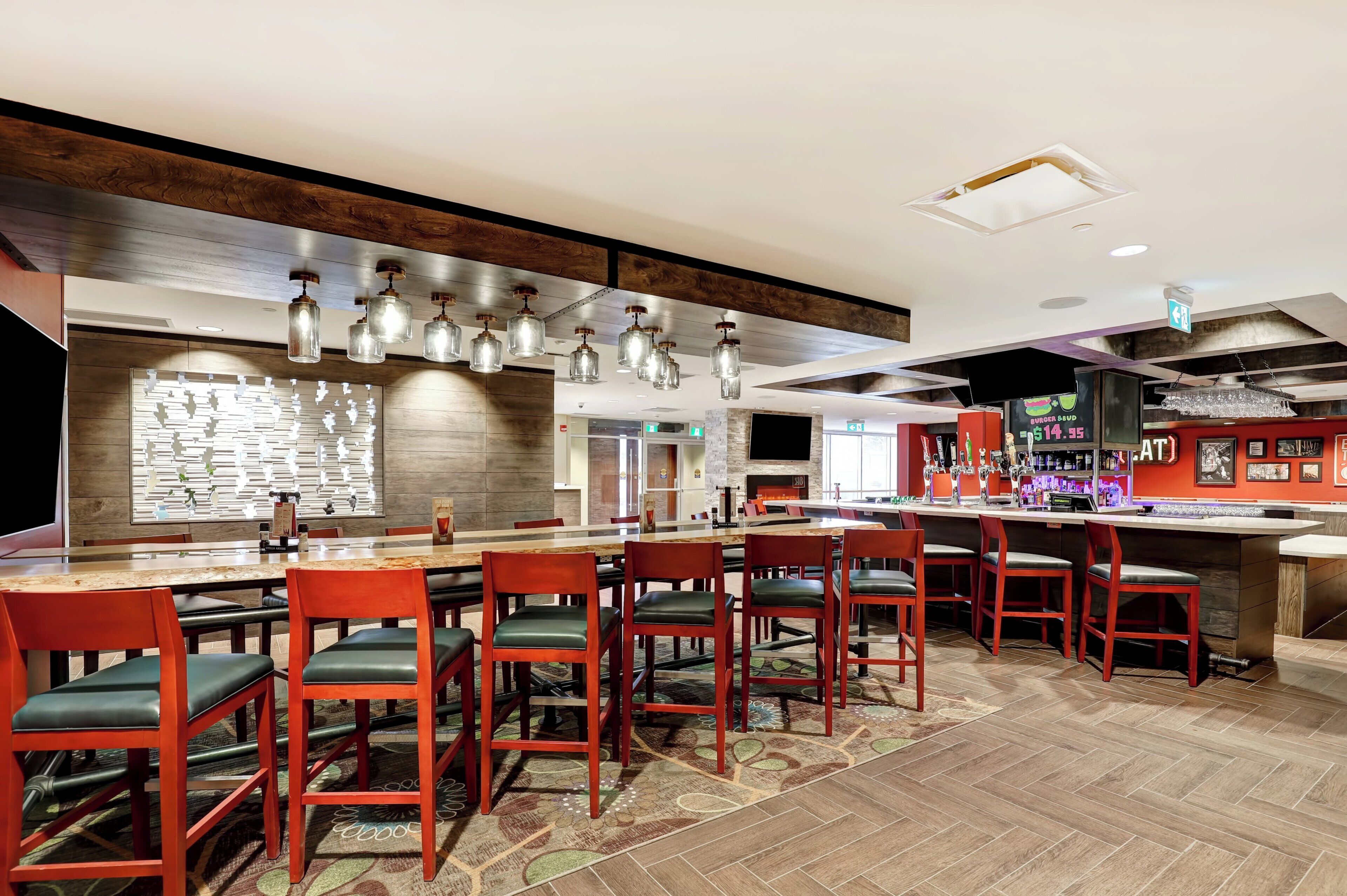 A restaurant with a long communal table, red chairs, hanging light fixtures, and a bar area.
