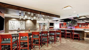 Un restaurante con una larga mesa común, sillas rojas, lámparas colgantes y una zona de bar.