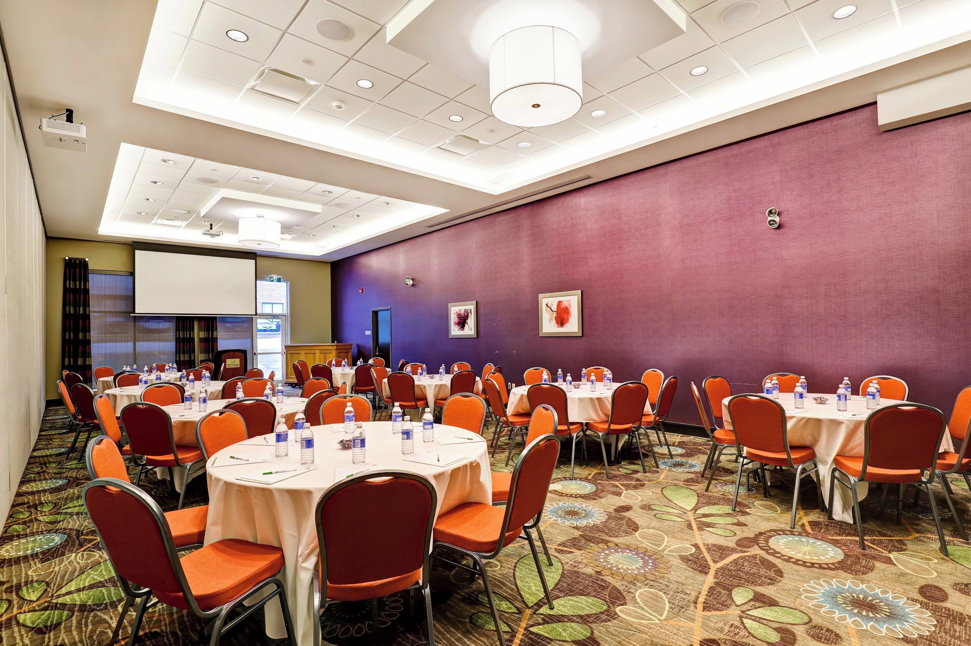 A conference room with multiple tables and chairs, a projector screen, and a large window.
