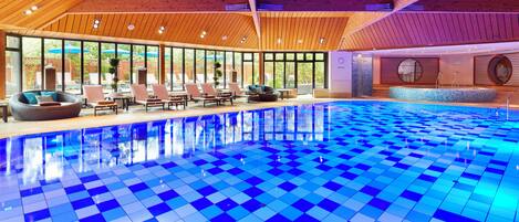Una amplia piscina interior con azulejos azules formando un diseño de cuadros, rodeada de sillones de descanso y un techo de madera.