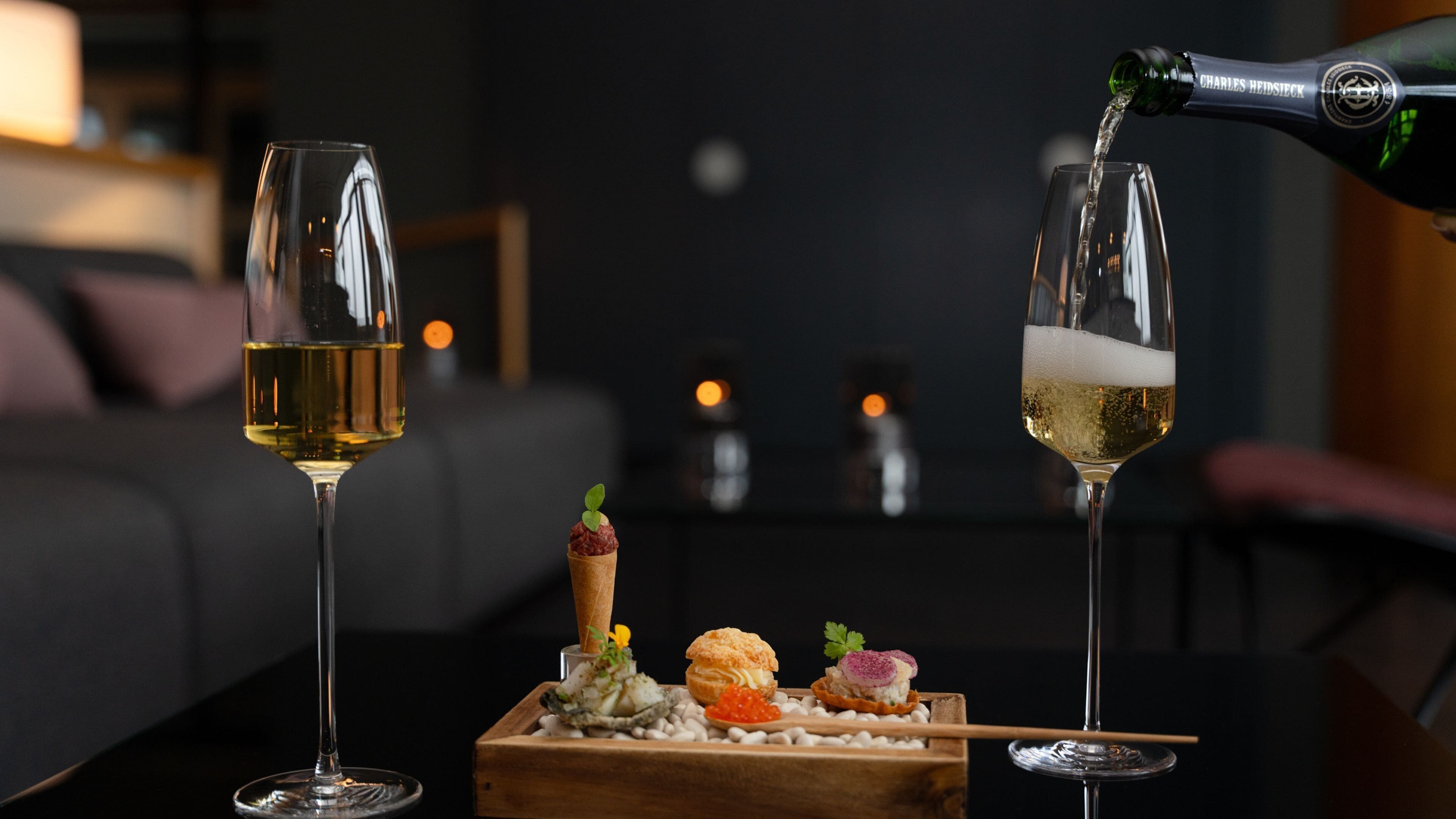 Two glasses of wine, one being filled, and a wooden box with food items on a table.