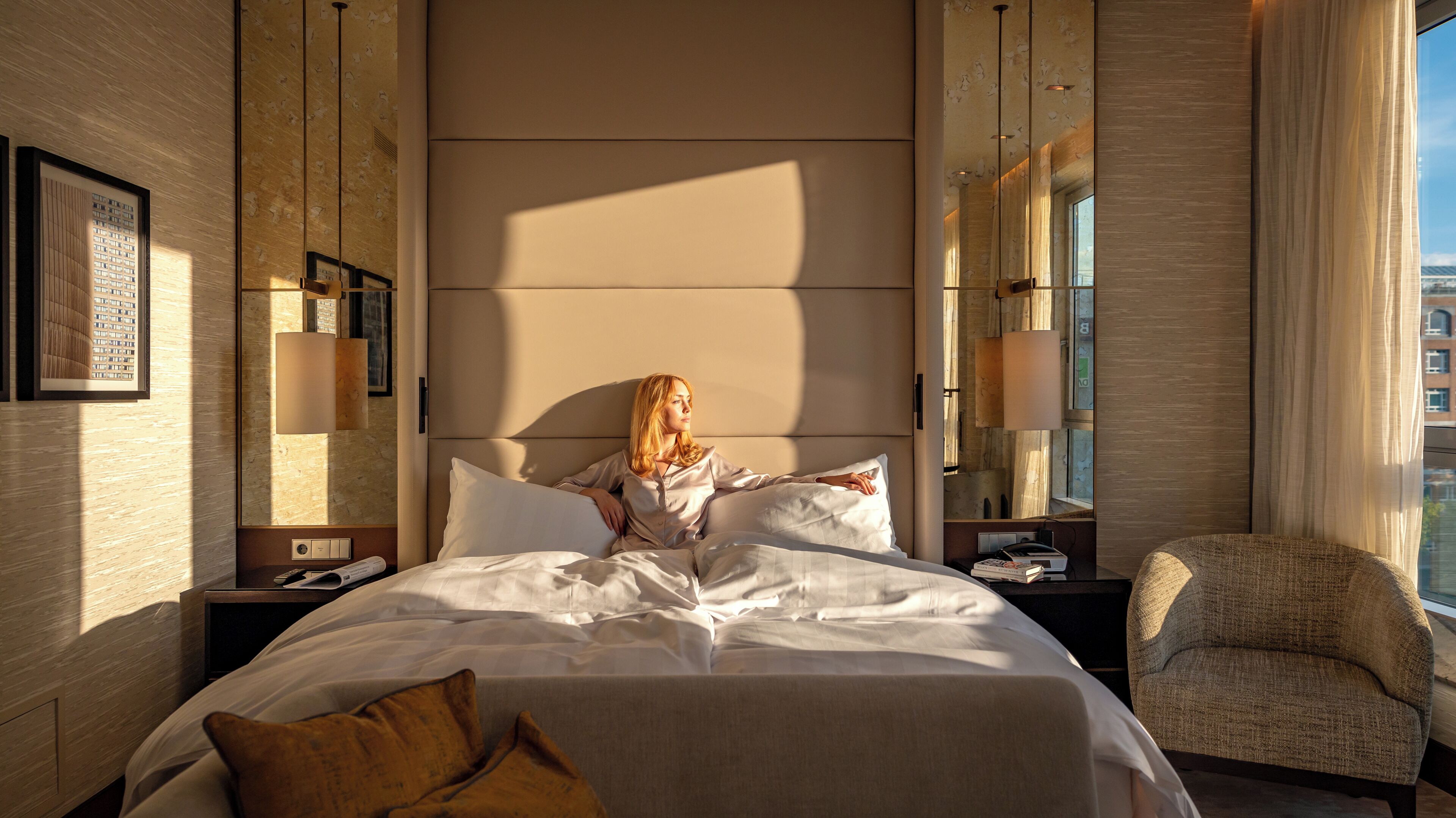 A hotel room with a large bed, a chair, a nightstand with a lamp, and a view of the city through the window.