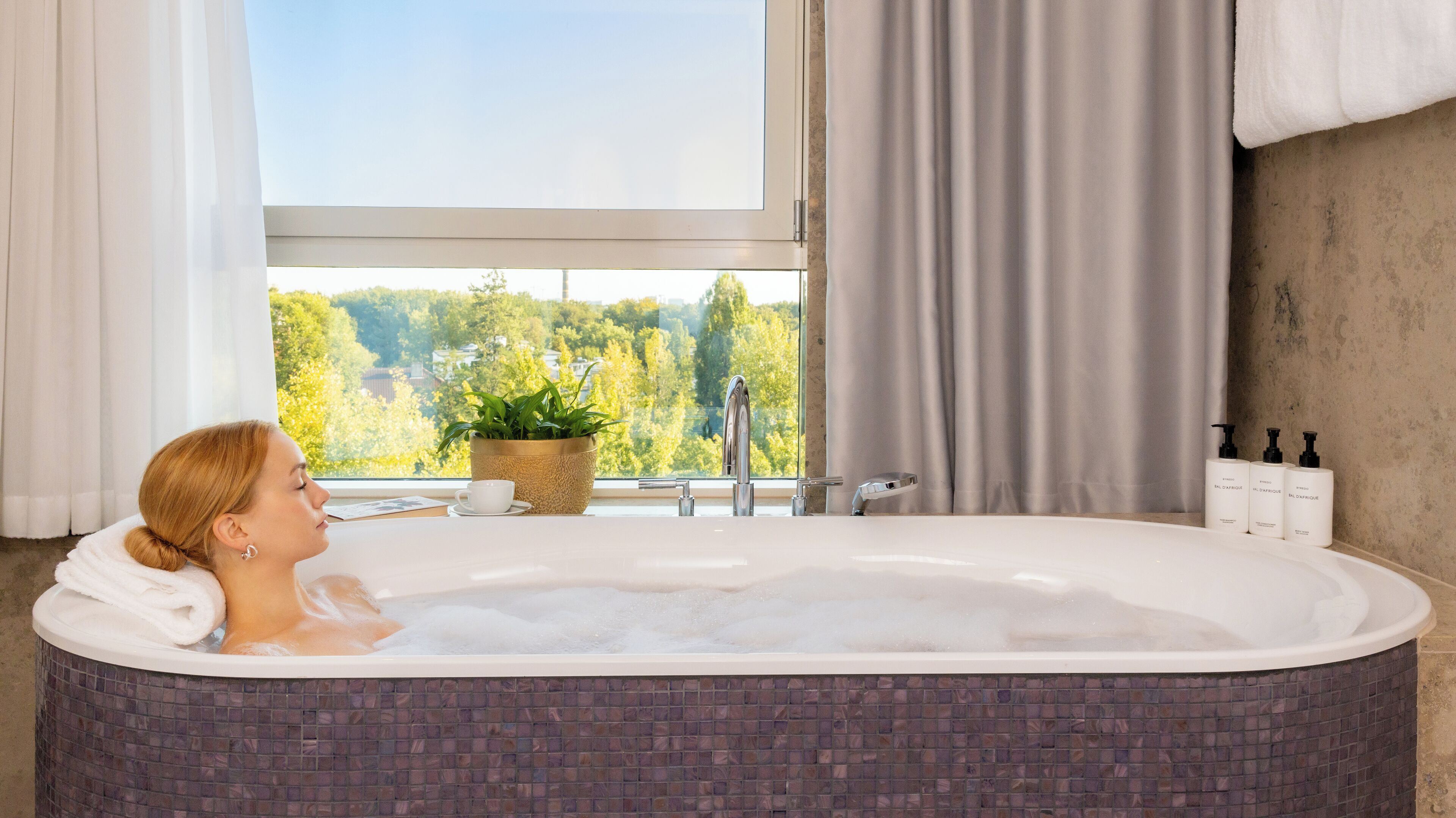 Eine Person entspannt in einer Badewanne mit Blick auf Bäume durch ein Fenster.