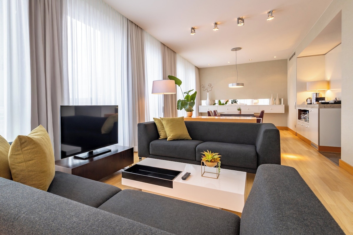 Una sala de estar moderna con un sofá gris, un televisor de pantalla plana y una mesa de centro con un jarrón. Se ve una zona de cocina con una mesa y sillas en el fondo.