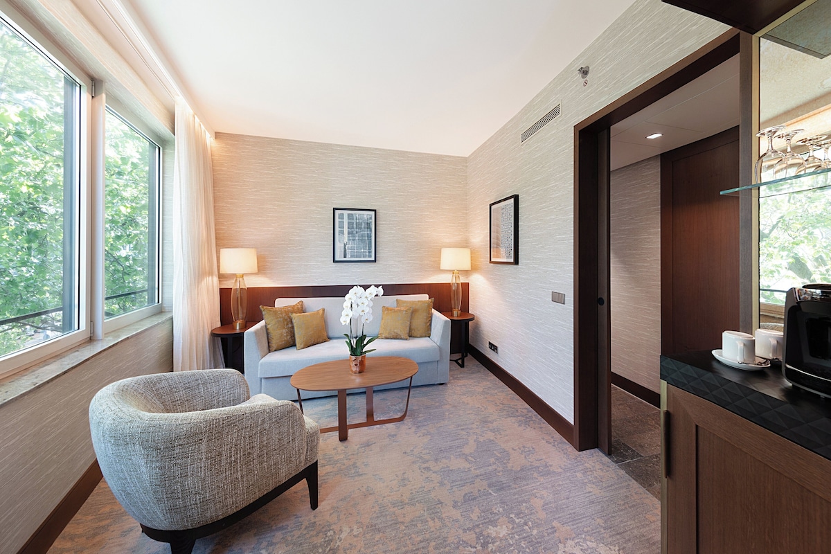 Una habitación de hotel moderna con sofá, sillón y mesa de centro, un rincón comedor con microondas, y ventanas amplias que ofrecen vistas a árboles.