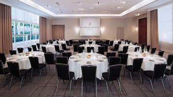 Un salón de conferencias con varias mesas redondas, sillas negras y una pantalla grande que muestra el nombre del hotel.