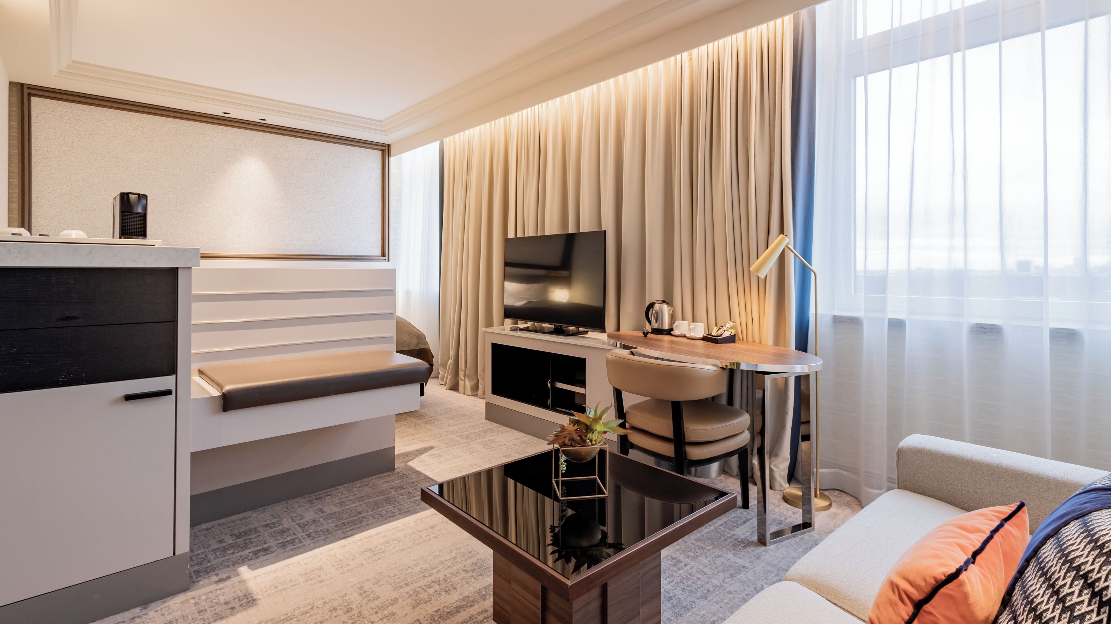 A modern hotel room with a flat-screen TV, a sofa, a coffee table, and a dining area with a lamp.