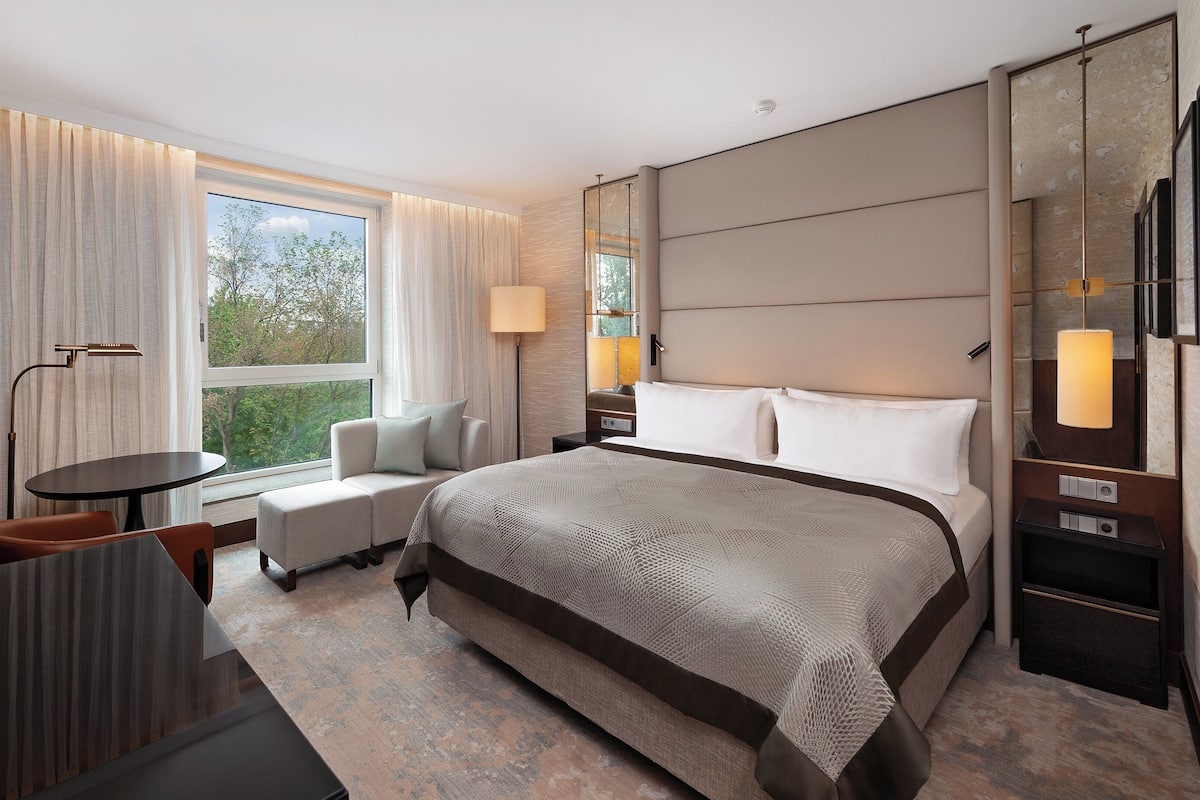 Moderni hotellihuone, jossa on suuri sänky, oleskelualue pöydällä ja tuoleilla, suuri ikkuna verhoilla sekä peili seinällä.