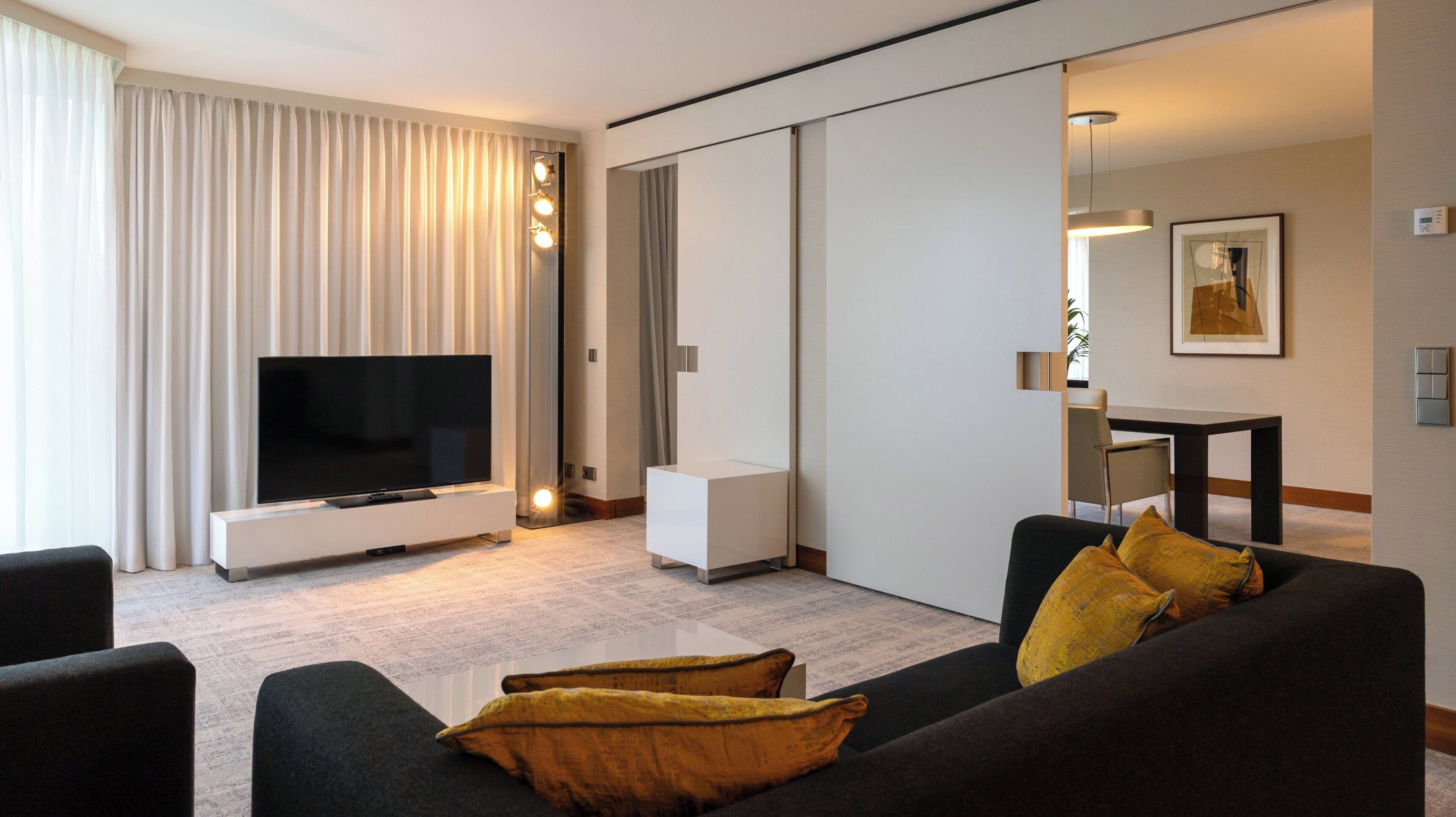 Ett modernt hotellrum med en platt-TV, en soffa med gula kuddar, ett soffbord och en skjutdörr som leder till badrummet.