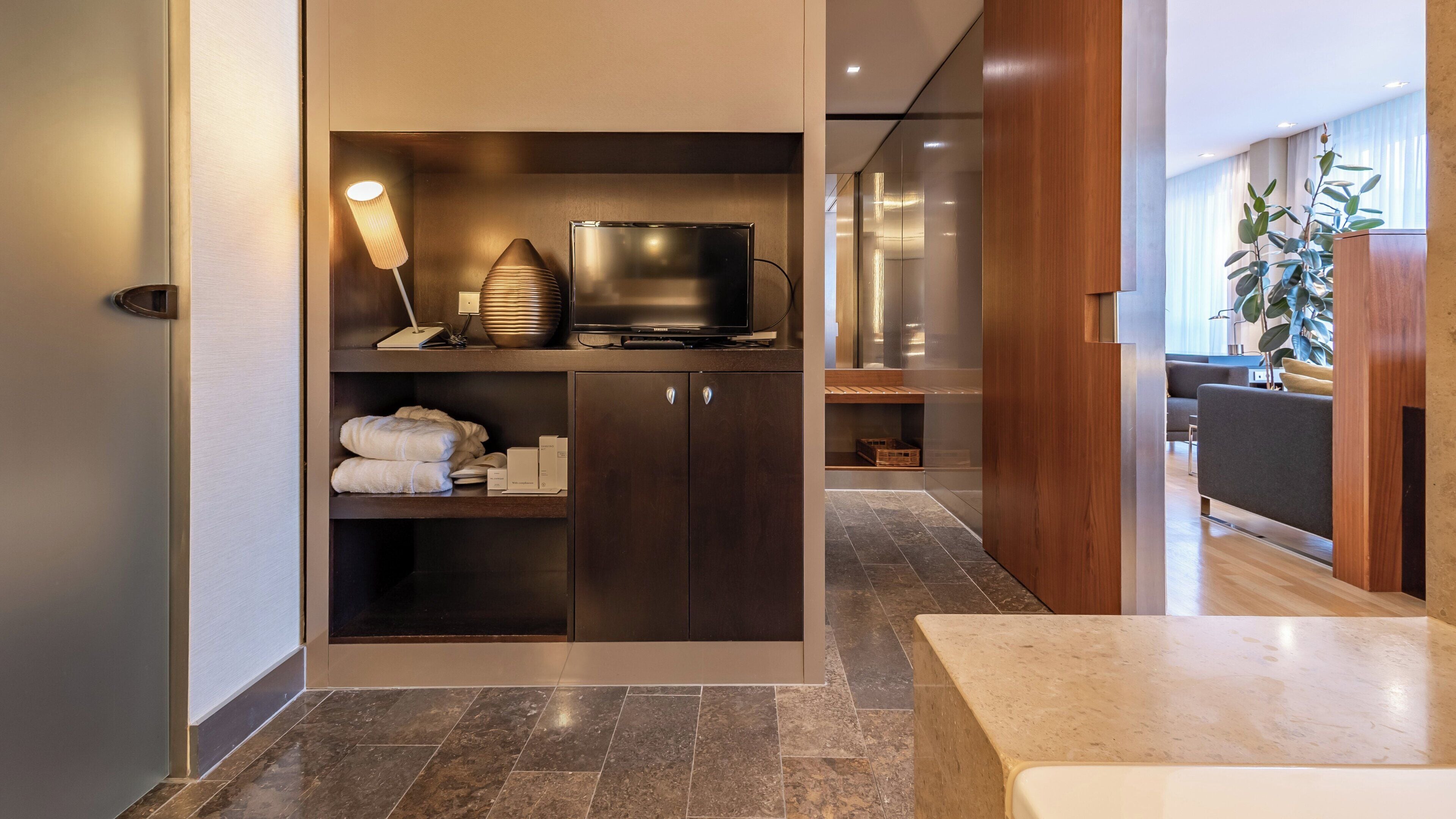 Una habitación de hotel moderna con una unidad de entretenimiento integrada, una zona de estar con sofá y un amplio ventanal con cortinas.