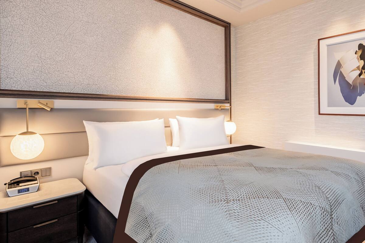 Moderni hotellihuone, jossa on suuri sänky, yöpöytä, jossa on kello, seinään kiinnitetty valaisin ja sängyn yläpuolella oleva taideteos.