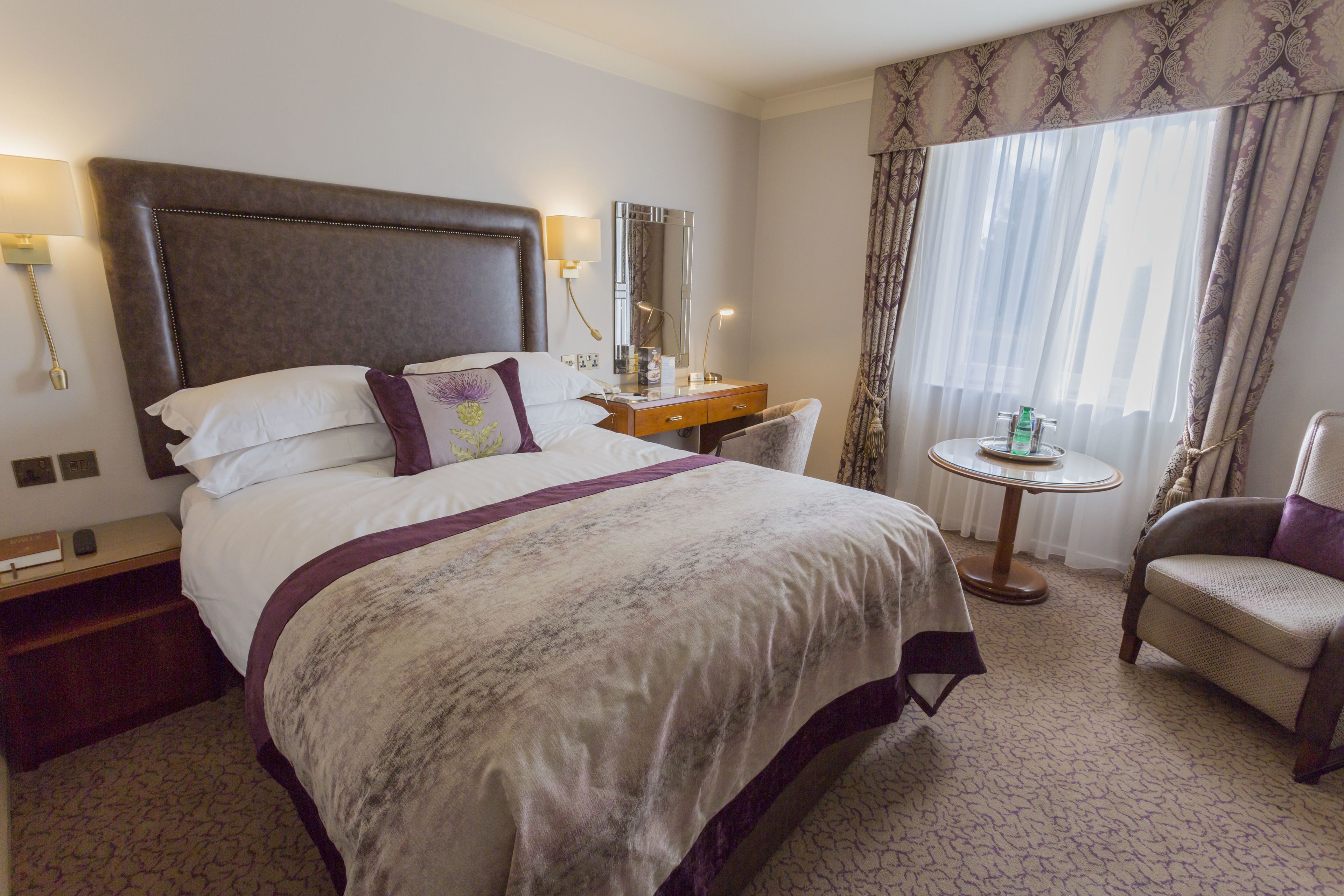 A hotel room with a large bed, a bedside table, a chair, a small round table, and a window with curtains.