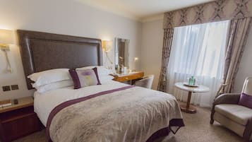 A hotel room with a large bed, a bedside table, a chair, a small round table, and a window with curtains.