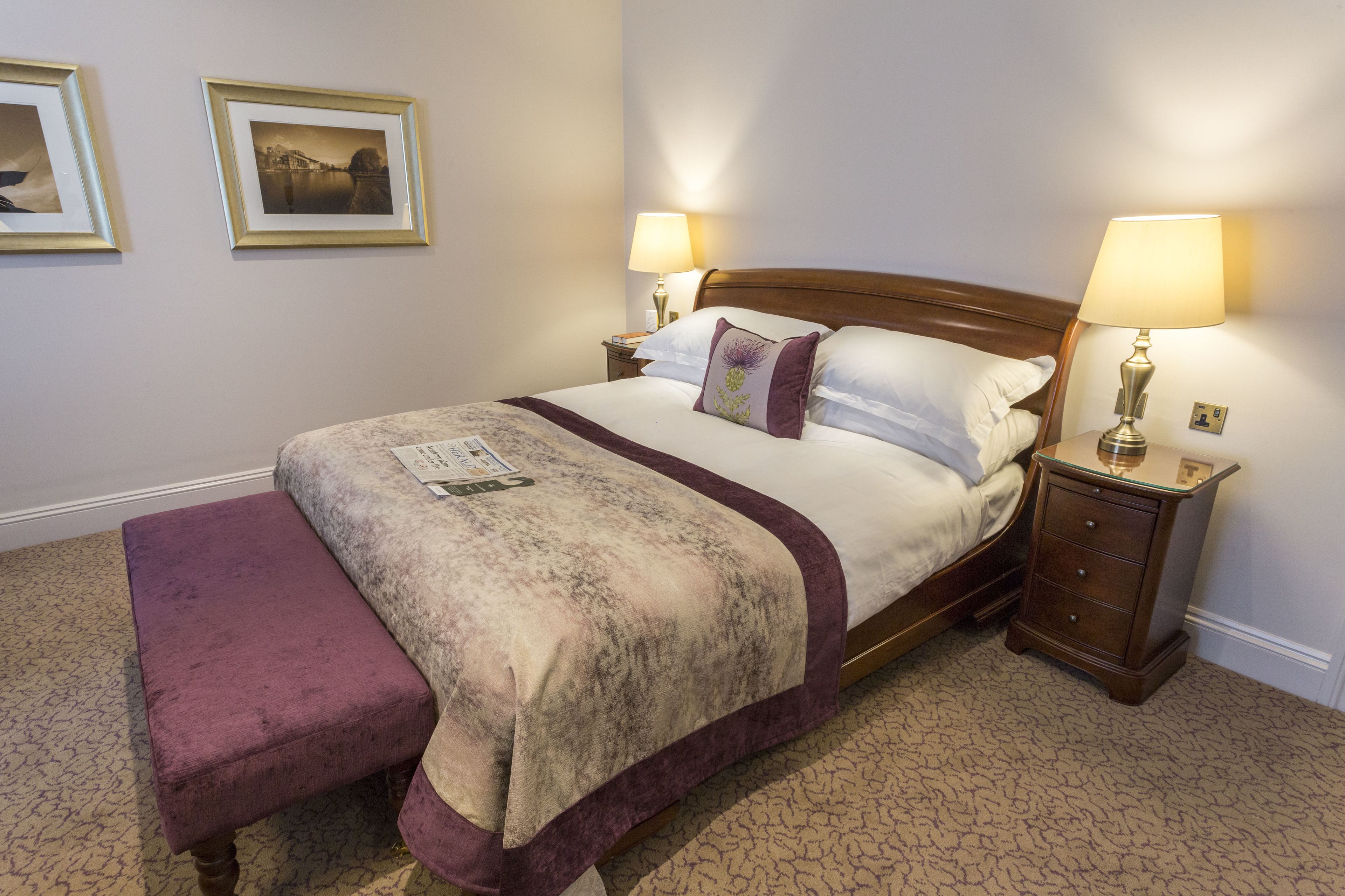 A neatly made bed with a headboard, two bedside tables with lamps, a bench, and two framed pictures on the wall.