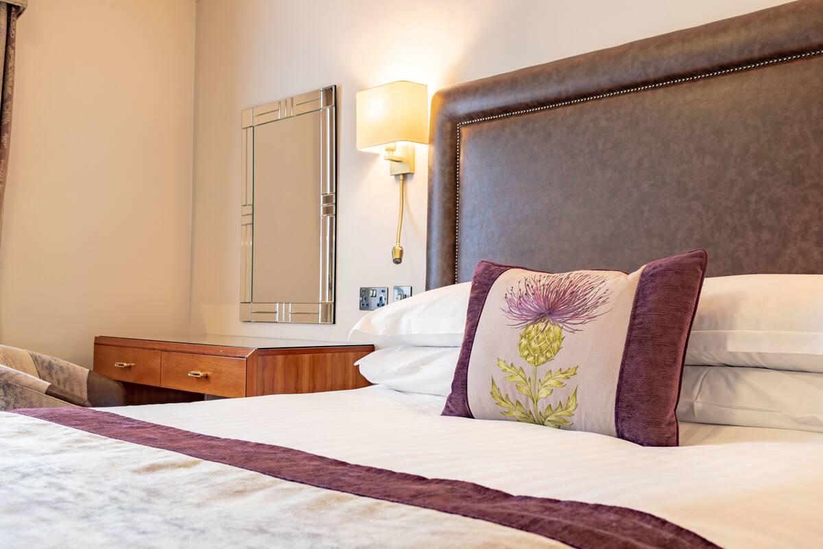Een netjes opgemaakt bed met hoofdbord, nachtkastje, lamp en spiegel aan de muur.