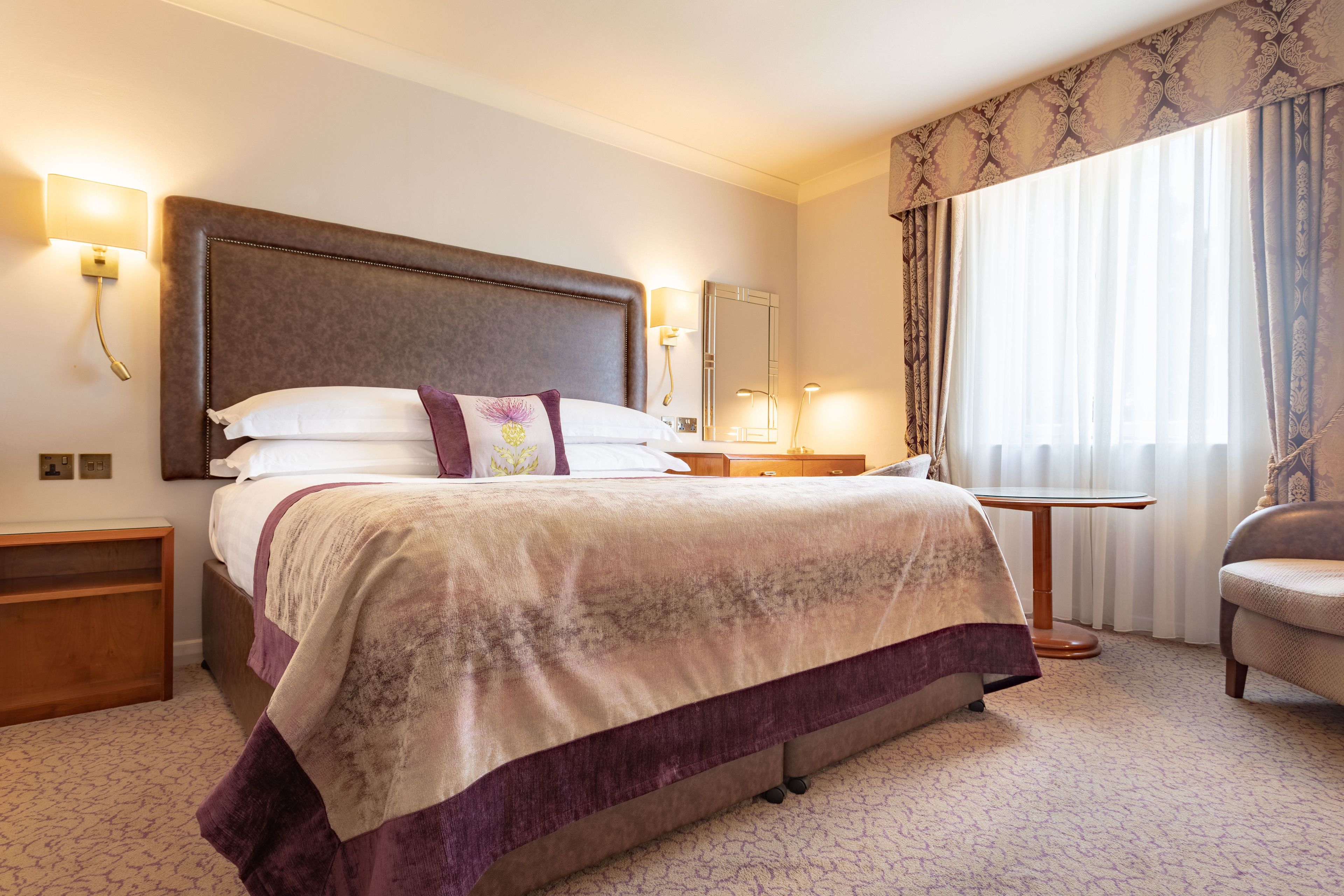 A hotel room with a large bed, a nightstand, a chair, a small table, and a window with curtains.