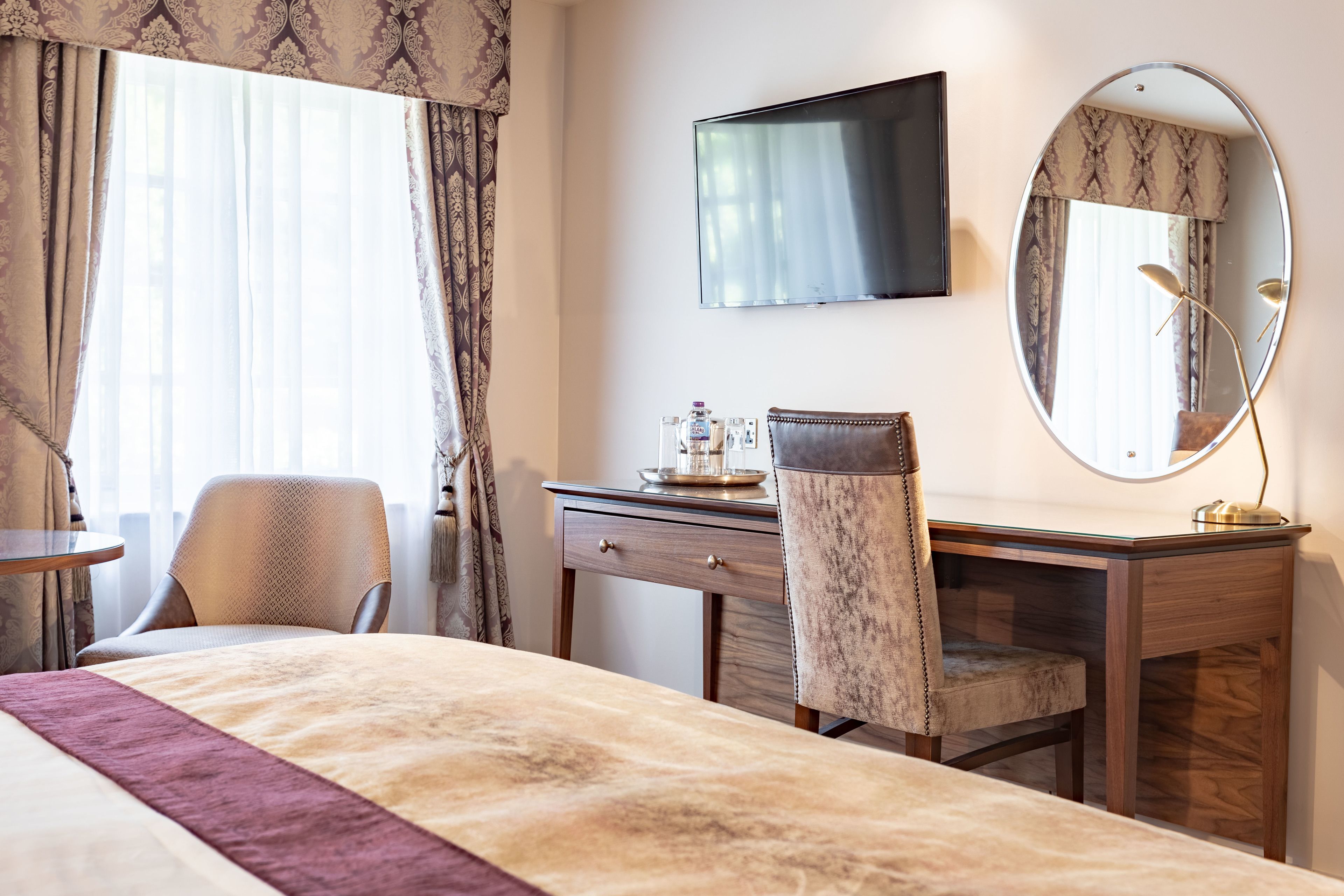 Een hotelkamer met een bed, een bureau, een stoel, een televisie, een spiegel en een raam met gordijnen.