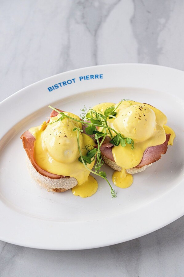 Un plato de Huevos Benedictinos con jamón y salsa holandesa, adornado con vegetales verdes, sobre un plato blanco con el texto 'BISTROT PIERRE'.