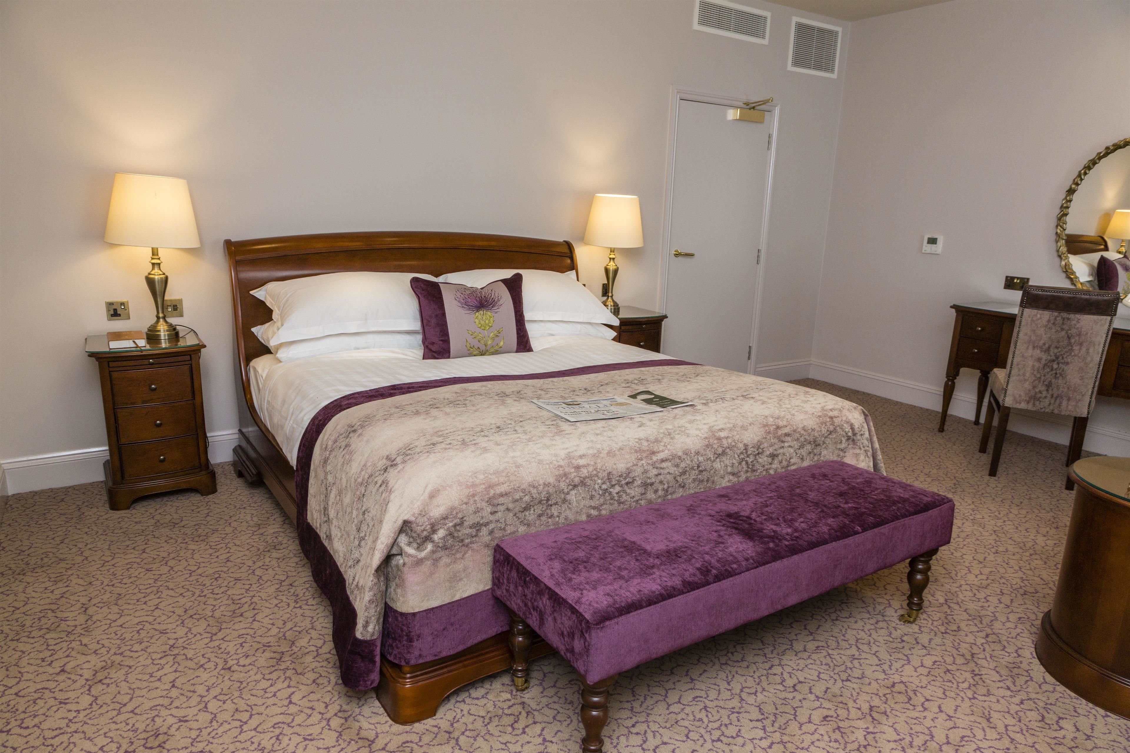 A neatly made bed with a wooden headboard, two bedside tables with lamps, a bench, a dresser, and a mirror on the wall.