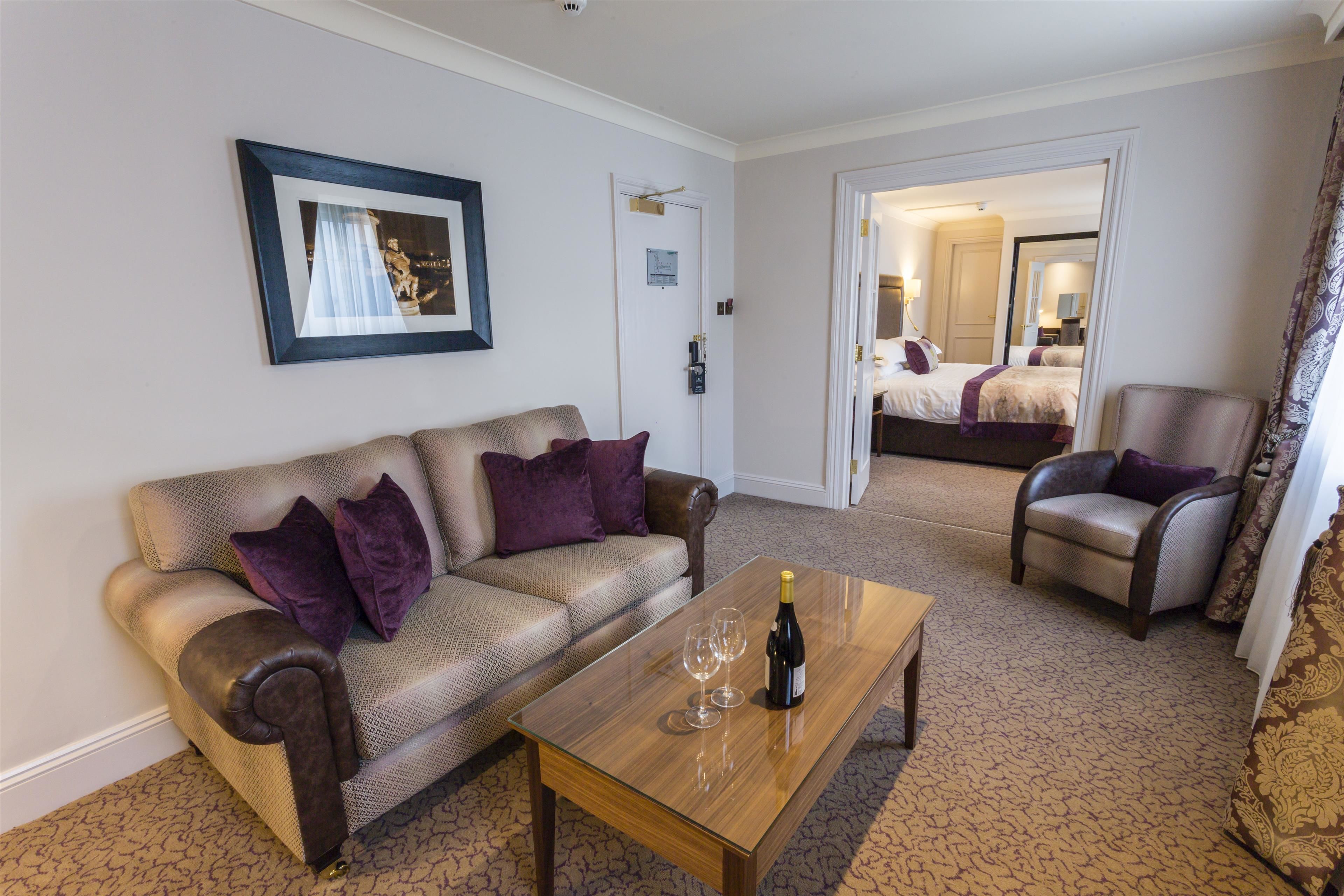 A hotel room with a leather sofa, a wooden coffee table with a bottle and glasses, a chair, and a bed in the background.